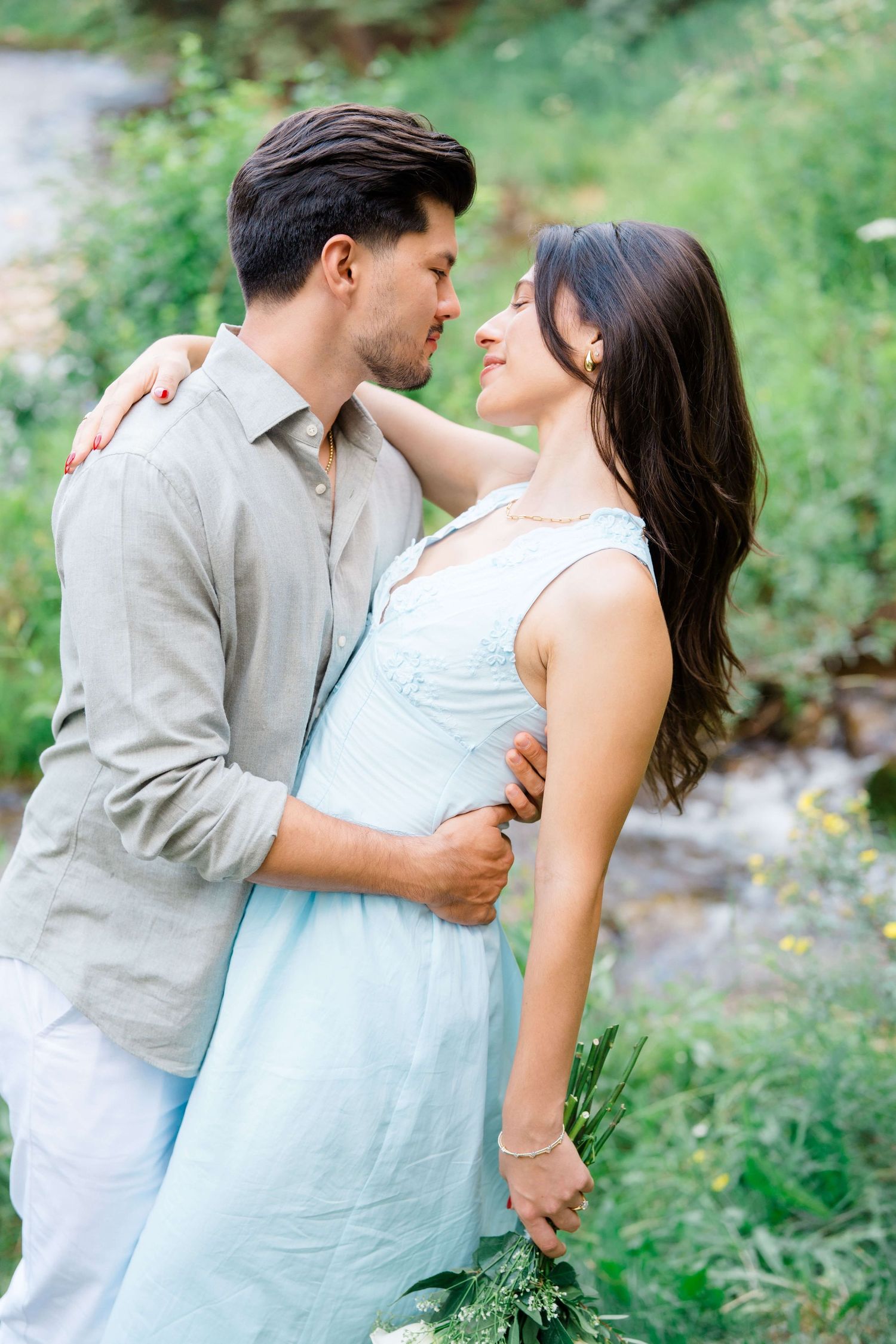 A couple embraces outdoors in a garden setting, with her wearing a light blue dress and him in a beige shirt after vail village proposal