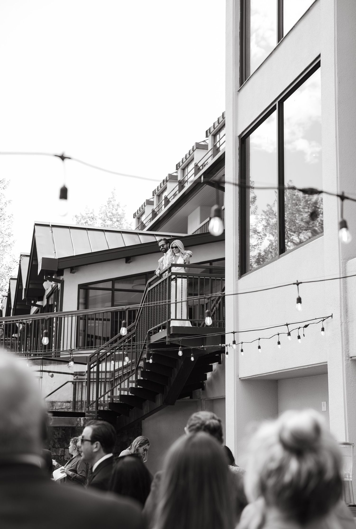 Black and white photo of guests gathered outside a multi-level venue with string lights and a staircase.