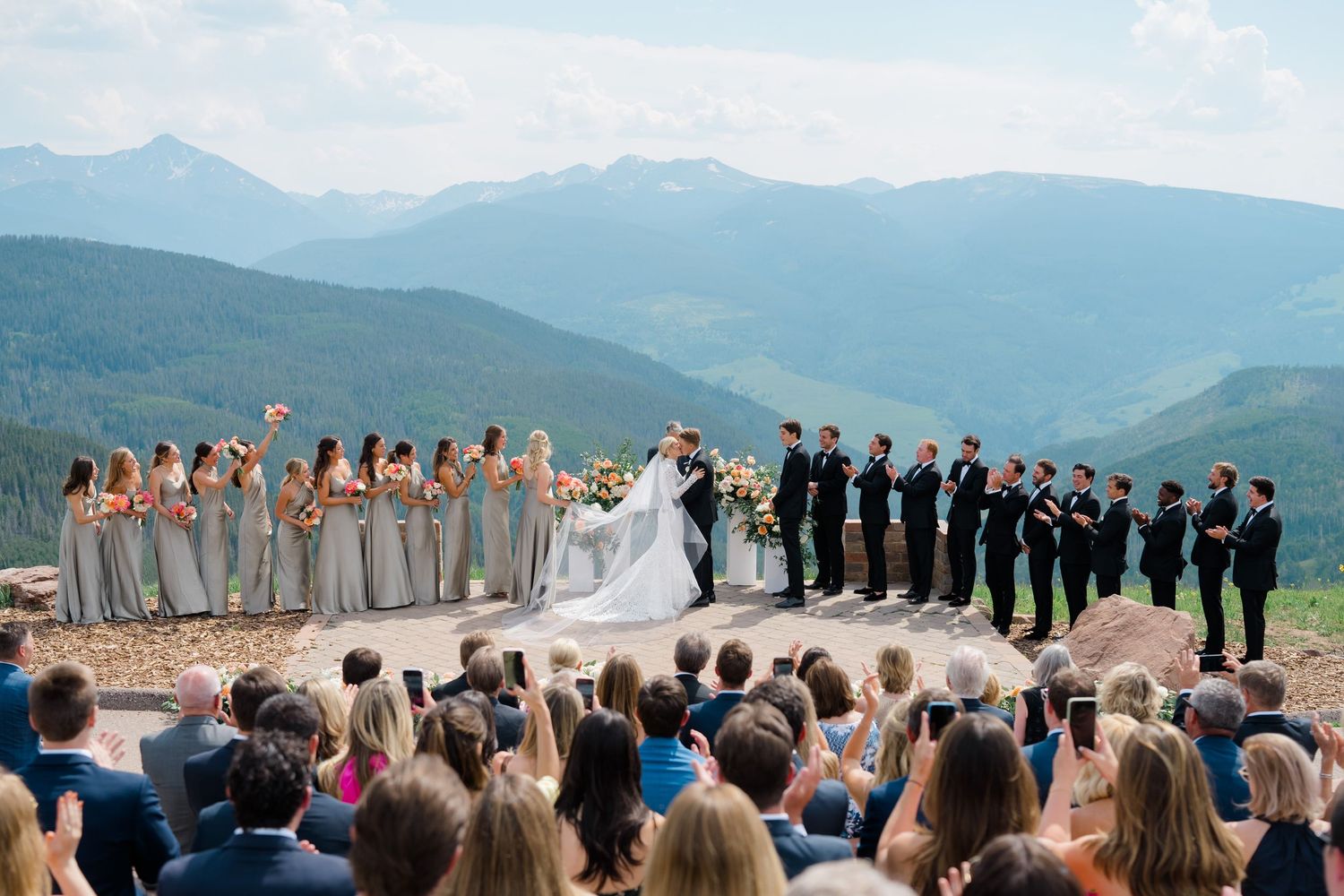 Mountain wedding ceremony with bride, groom, and wedding party standing before seated guests with scenic mountain views.