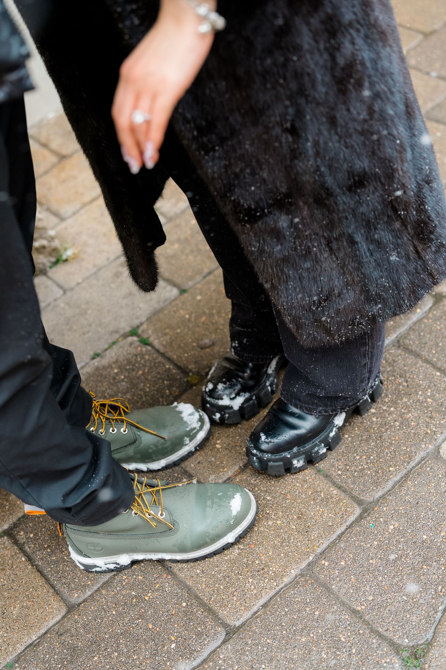 Two people standing on brick pavement in winter boots, one wearing green lace-up boots and the other black patent chunky boots.