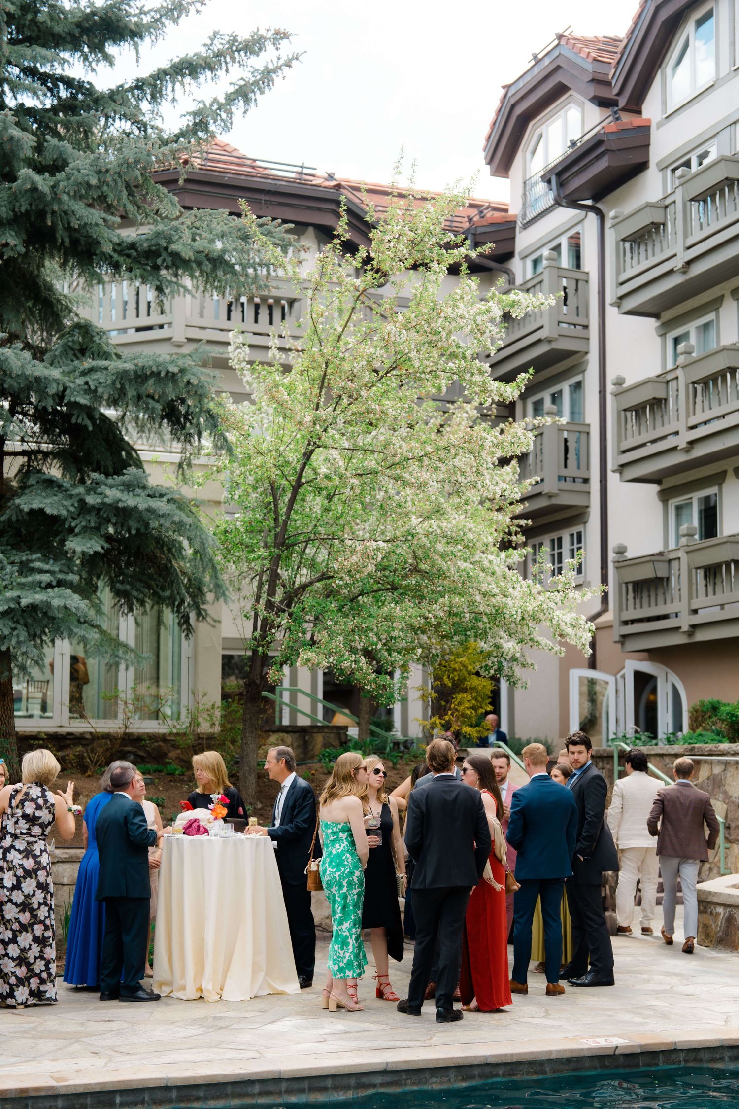 Sonnenalp Vail Hotel Elegant outdoor cocktail reception with guests in formal attire gathered near a blooming tree by a luxury hotel pool.