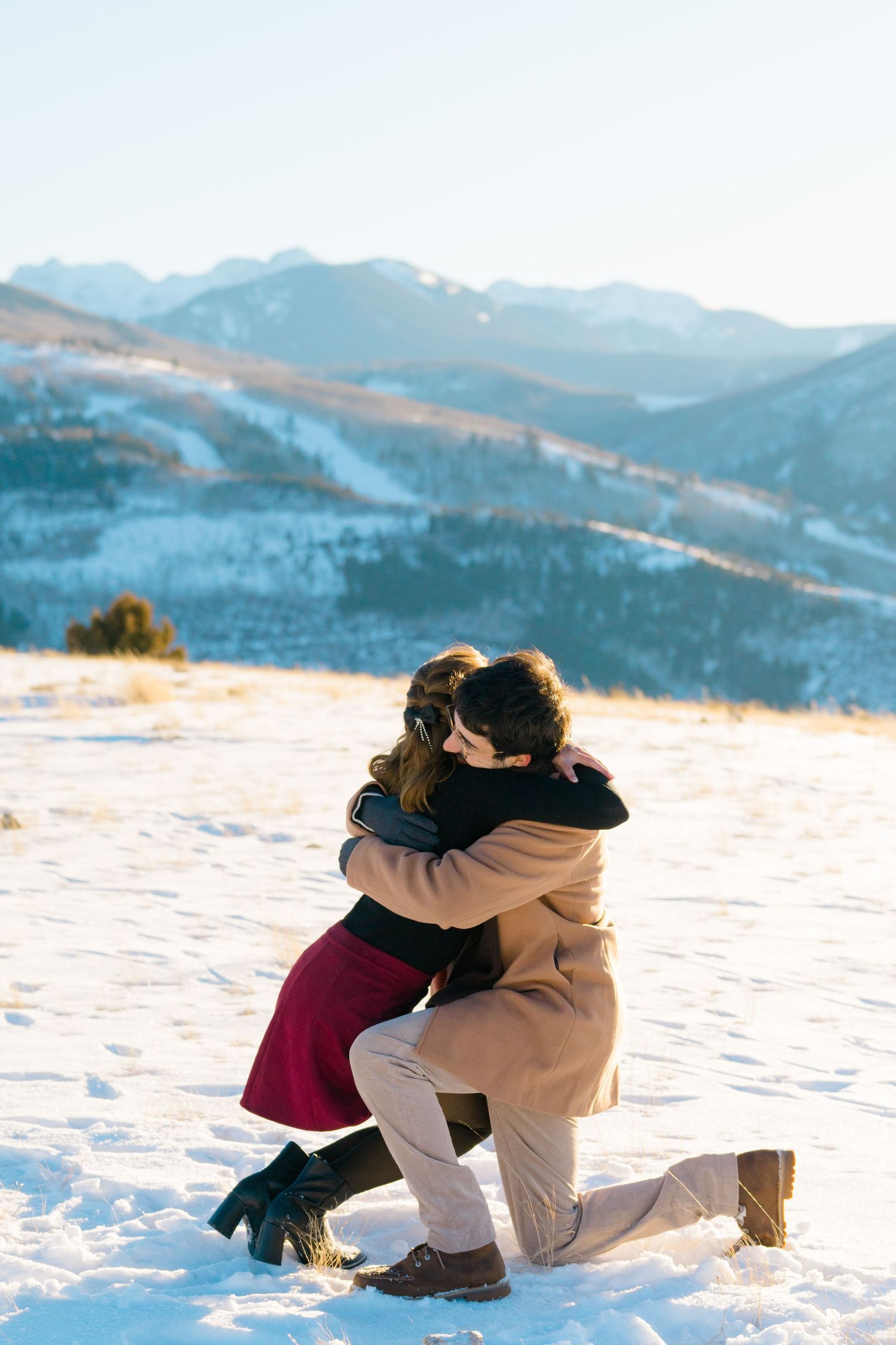 A couple sits together in the snow with scenic mountain views in the background during winter.