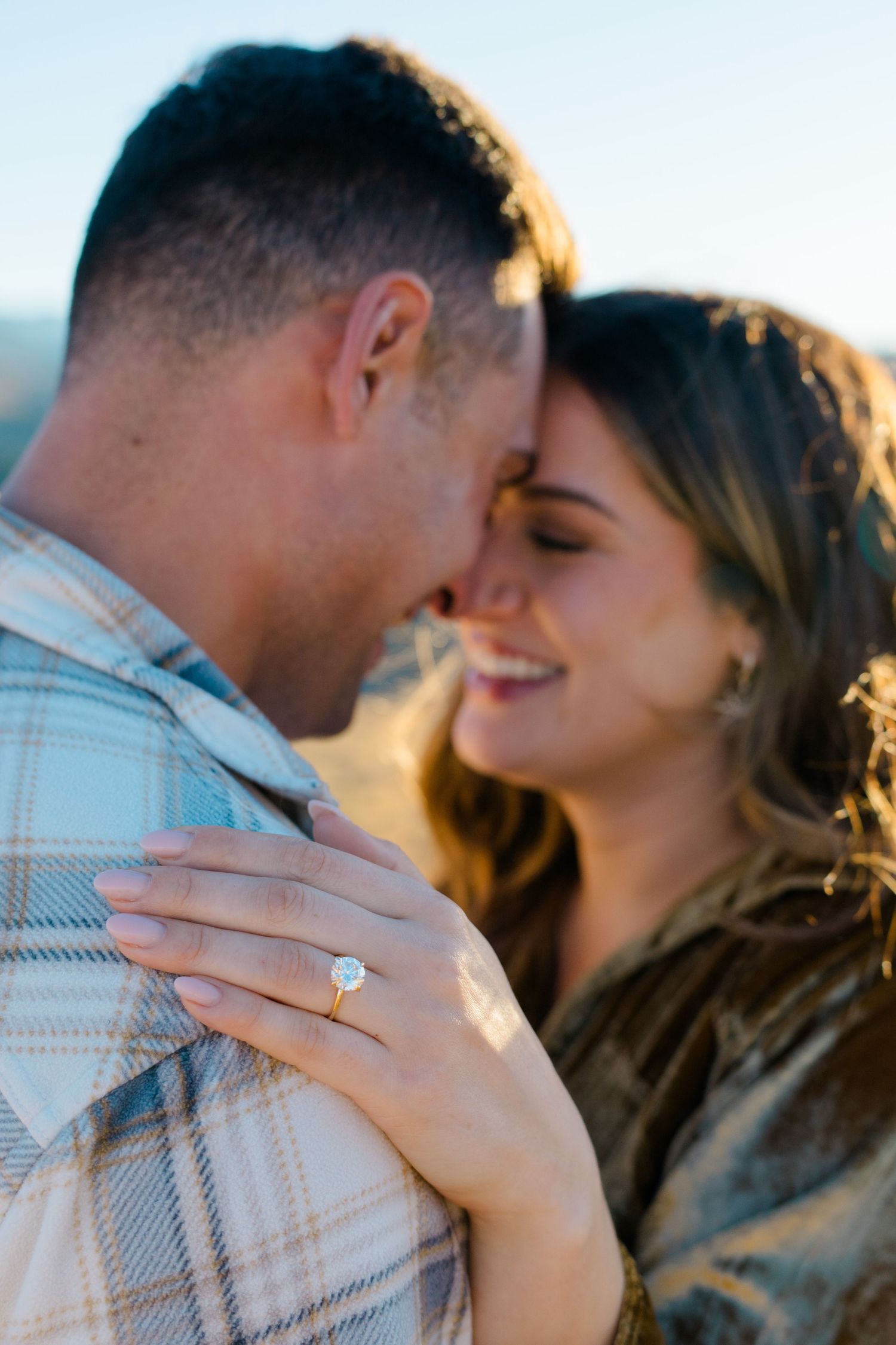 A romantic couple sharing an intimate moment while showing off a new engagement ring in warm sunlight.