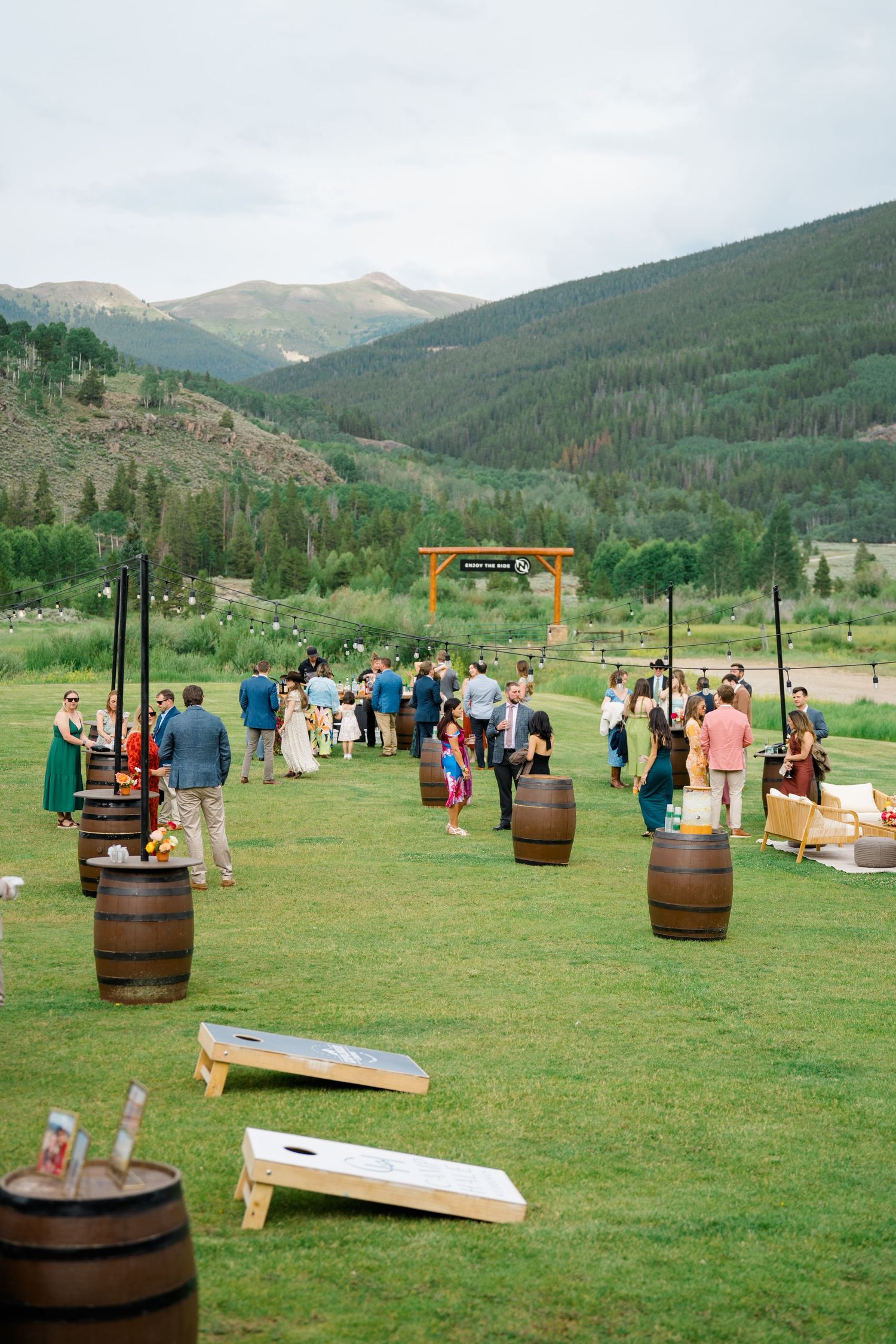 Outdoor wedding reception area with wooden barrels and cornhole games set up on mountain meadow.