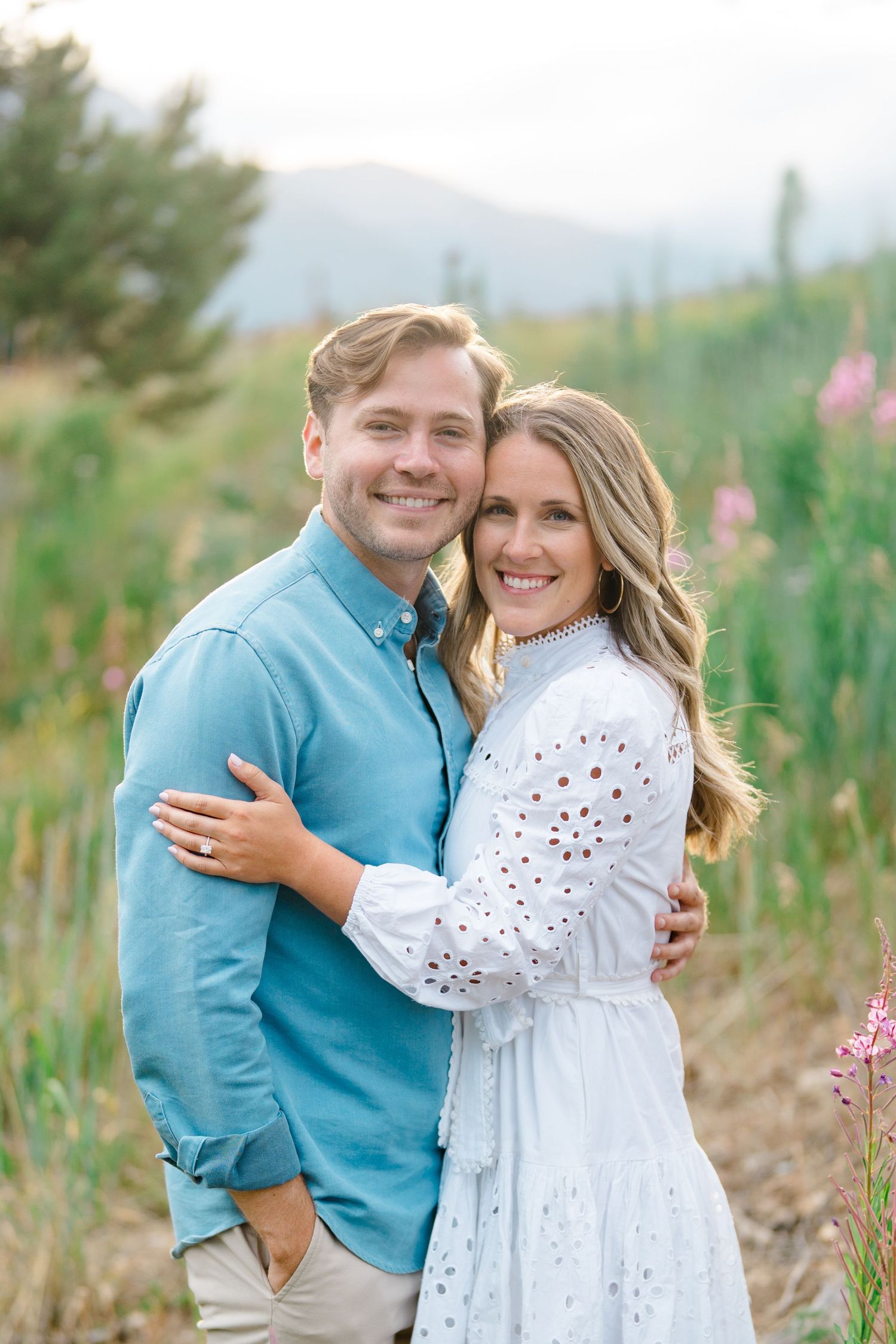 A couple poses together in a field of wildflowers with mountains visible in the background.