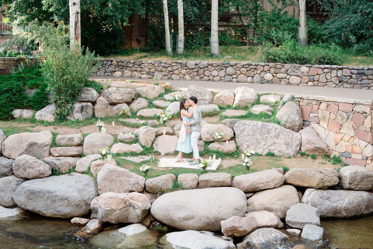 A figure in white stands on natural stone steps leading down to a tranquil stream surrounded by lush greenery.