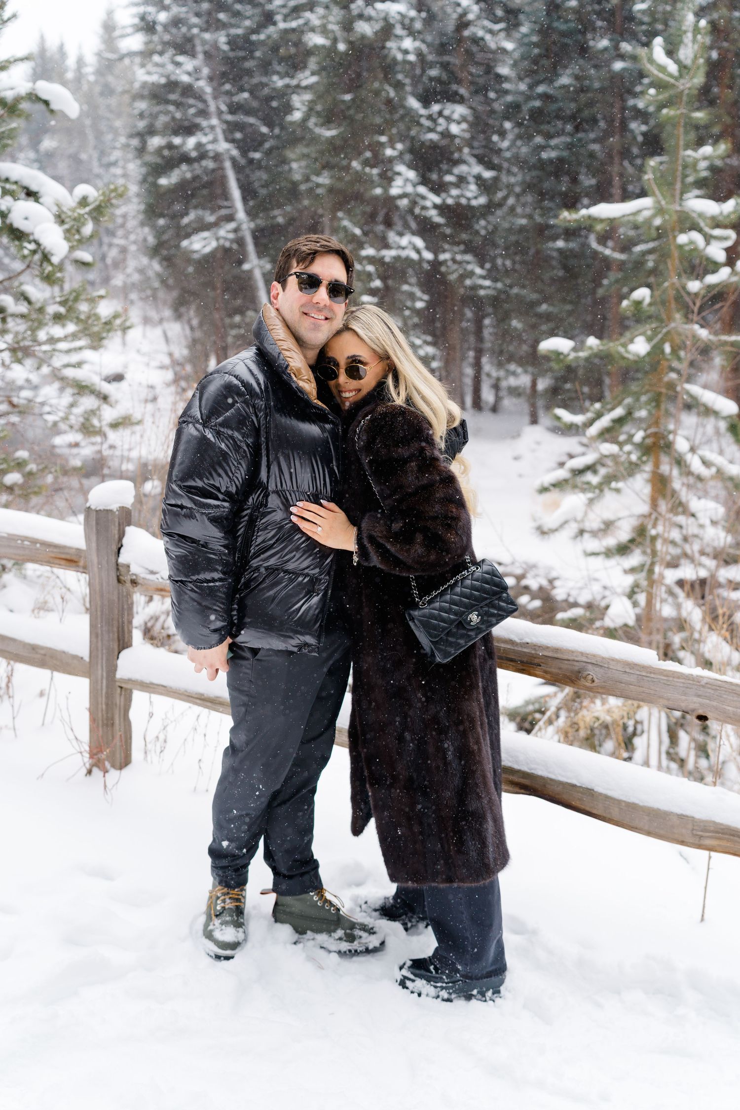 Couple embracing in snowy winter forest, wearing black puffer jacket and dark fur coat with sunglasses.