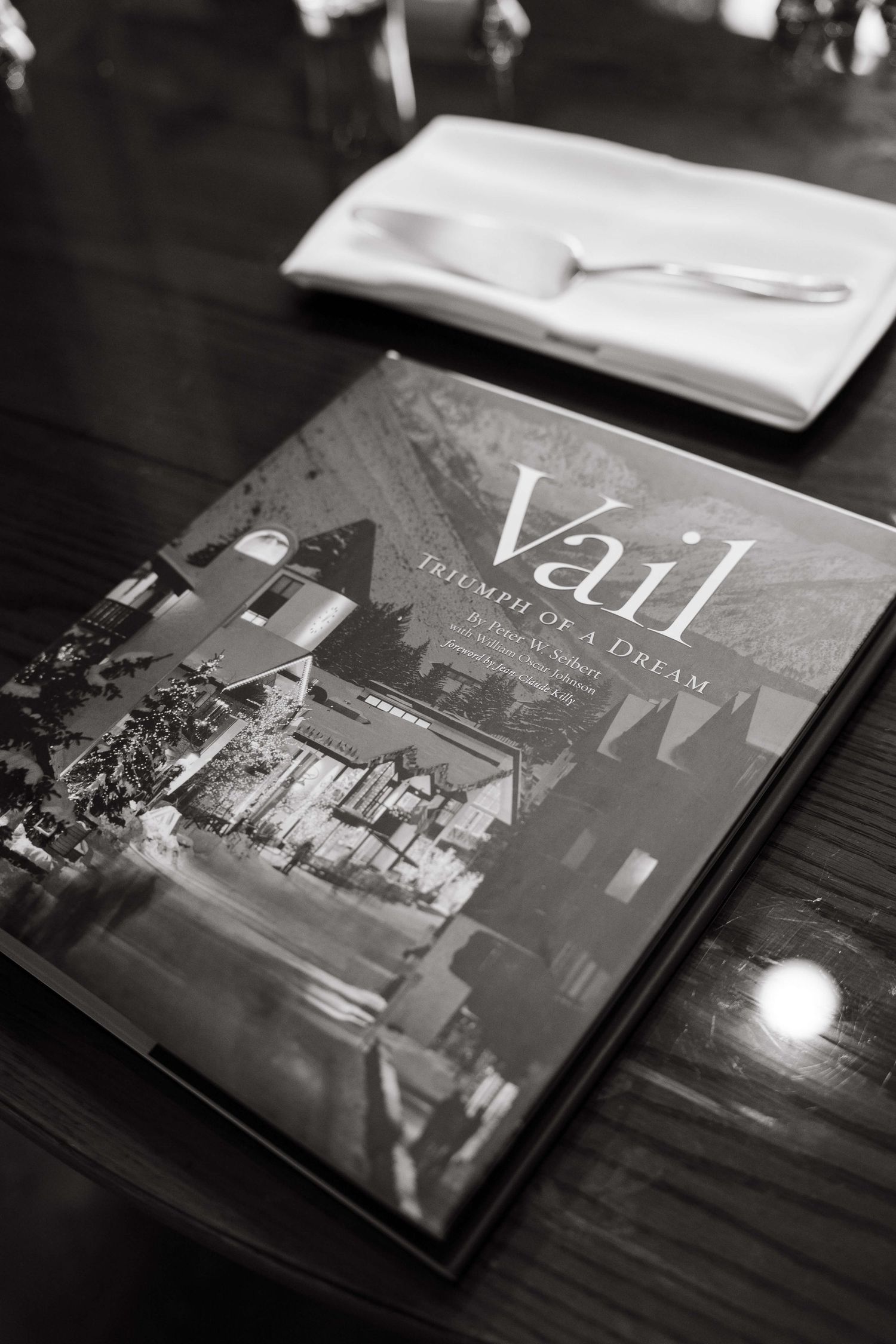Black and white photo of Vail: Triumph of a Dream book on a wooden table with silverware in background.