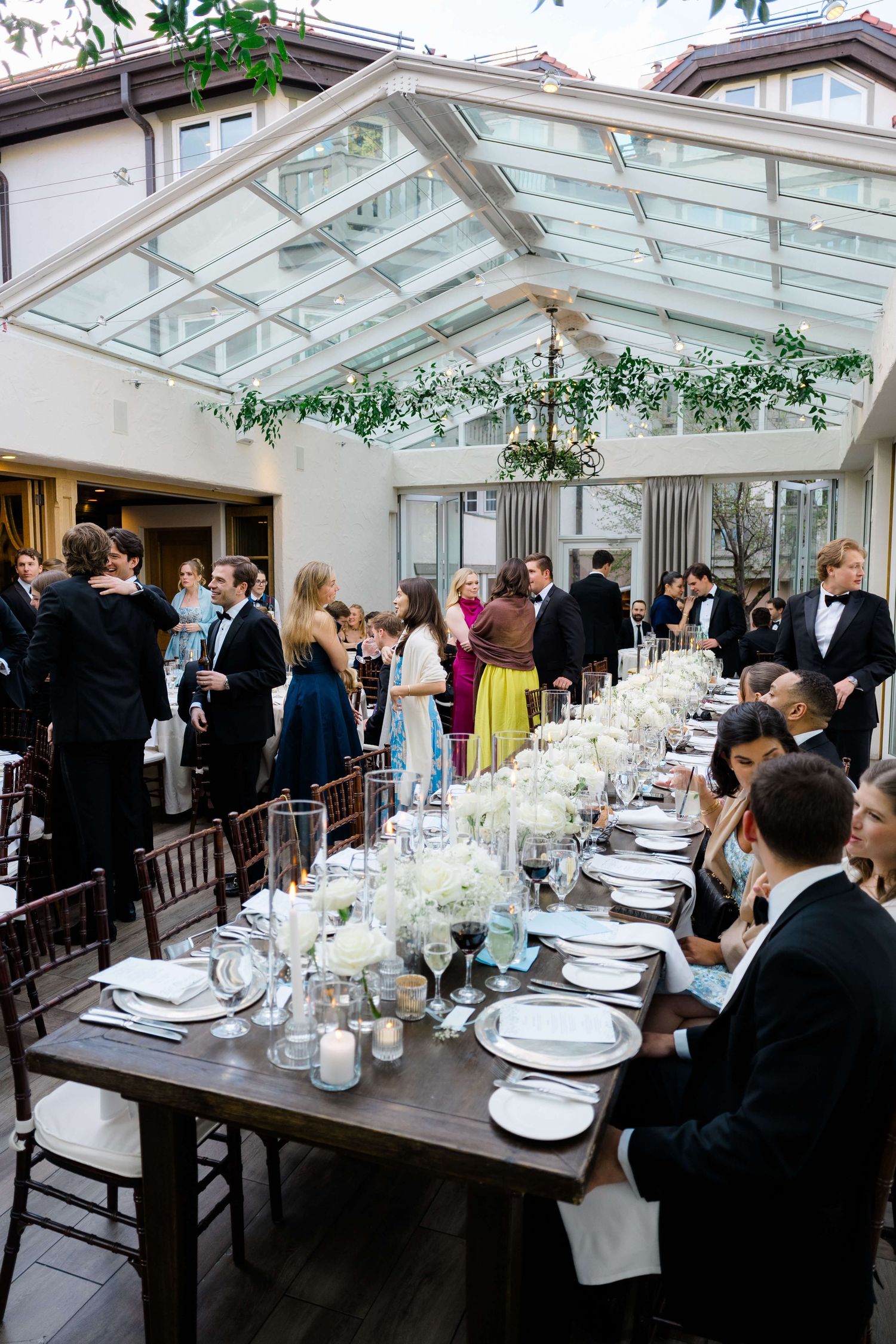 Elegant wedding reception in a glass-roofed venue with long dining tables, white floral centerpieces, and formally dressed guests.
