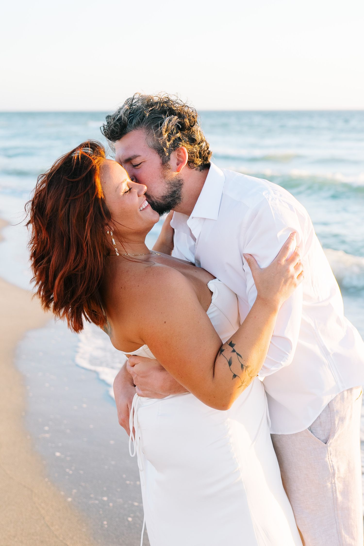A romantic couple shares intimate kisses on a sunny beach with ocean waves in the background.
