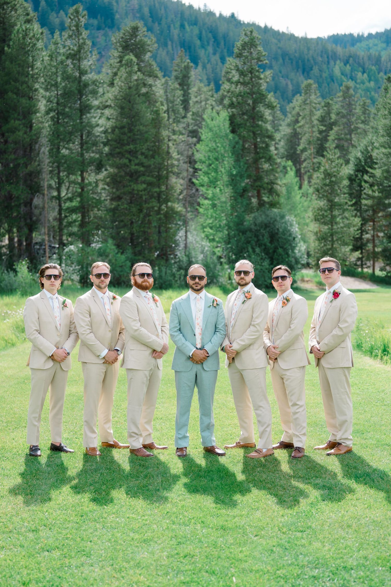 Groomsmen in light tan suits and sunglasses pose together on grass with mountain forest backdrop.