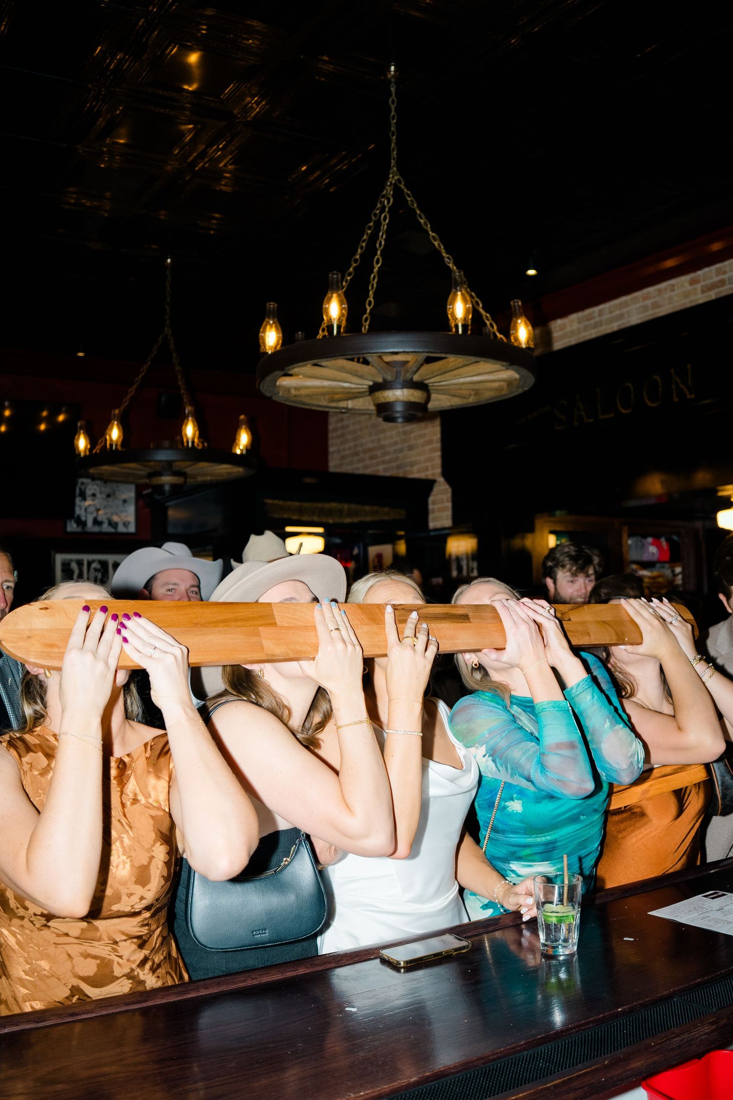 A row of people lying face down on a bar counter in a dimly lit establishment with hanging lights above.