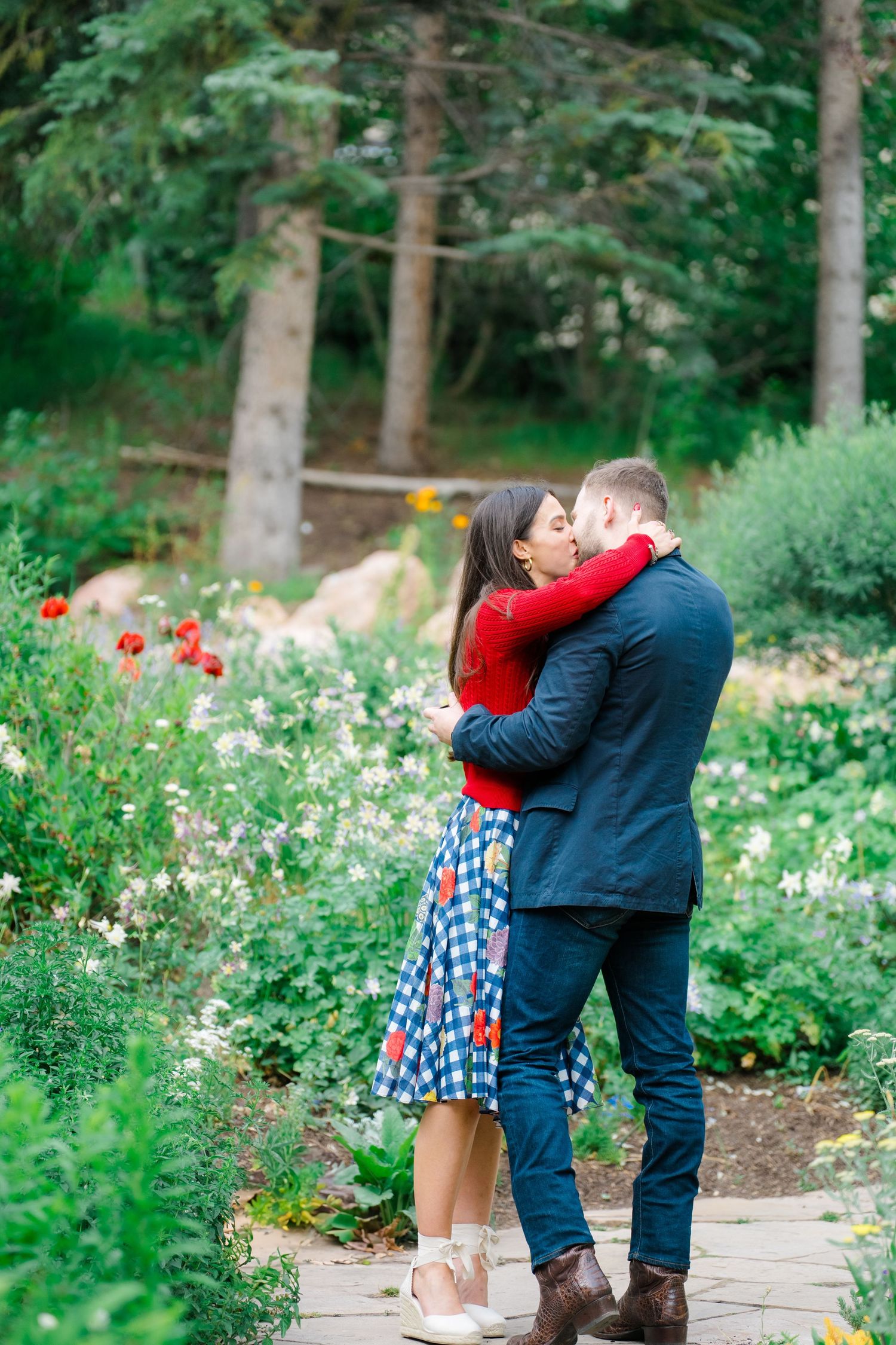 A couple embraces romantically in a garden path surrounded by blooming flowers and lush greenery.