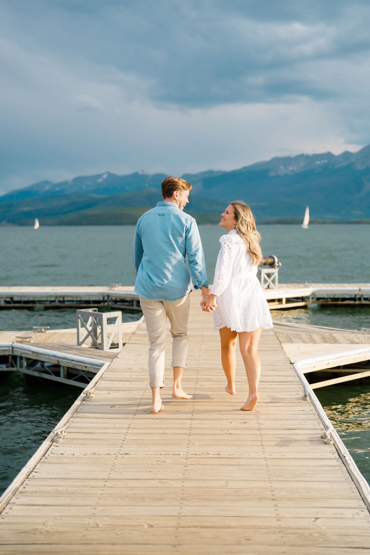 Two people walk hand in hand down a wooden dock over a lake with mountains in the distance.