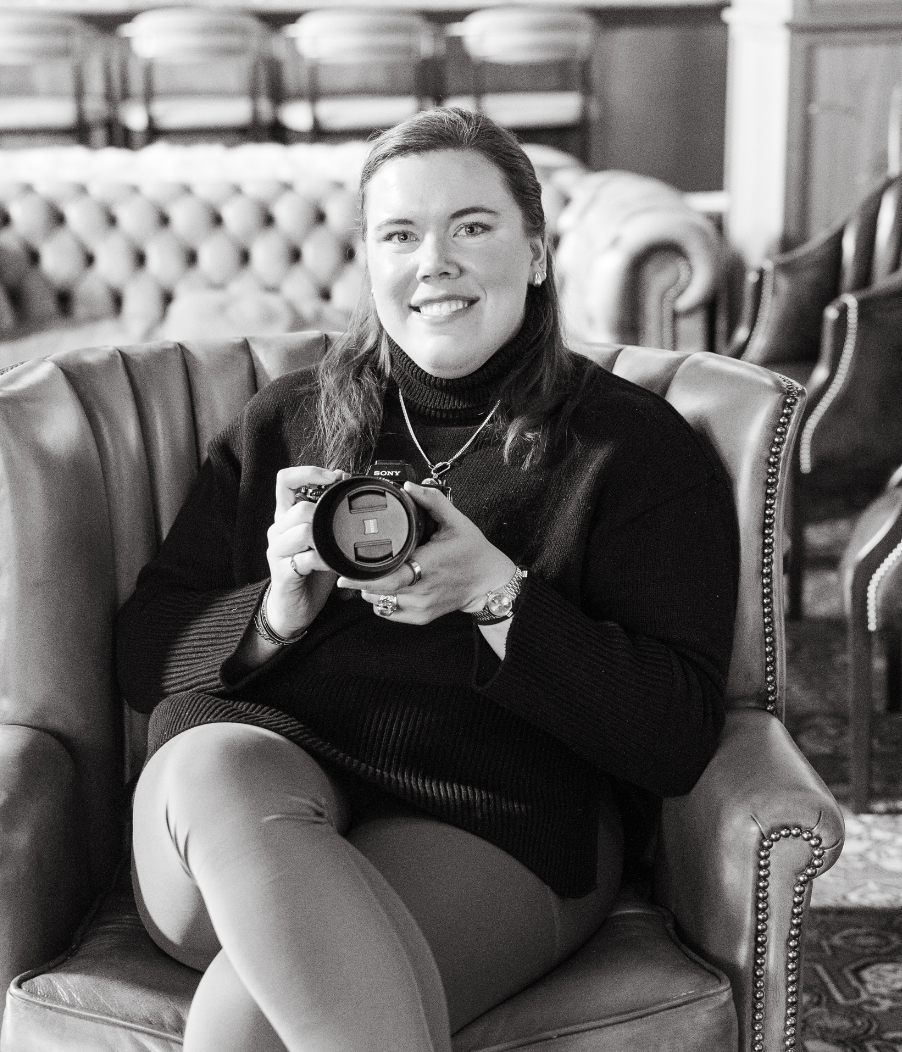 A photographer in black attire sits comfortably on a leather couch while holding a vintage camera.