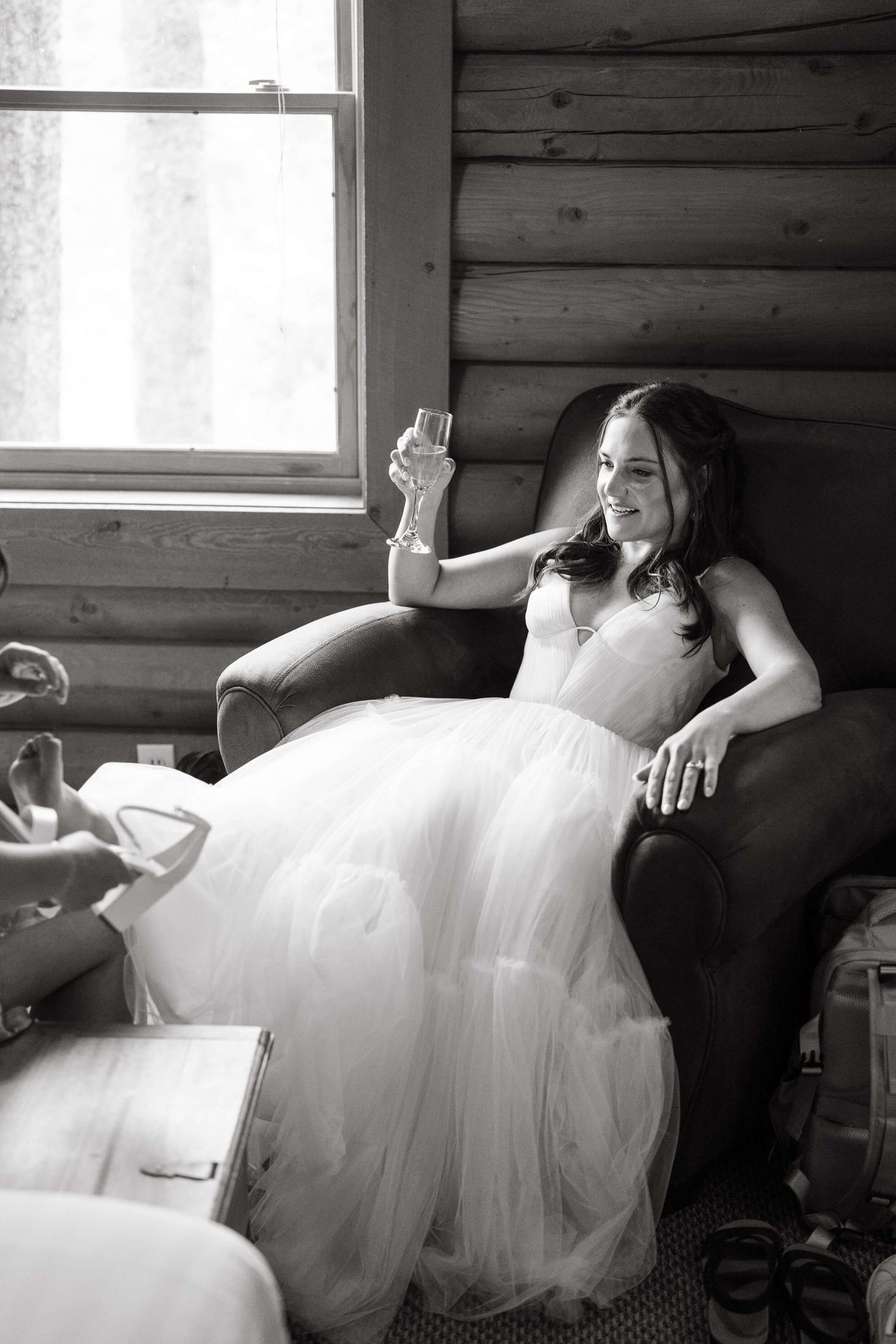 Black and white photo of a bride in a flowing tulle gown relaxing in a rustic log cabin setting at Camp Hale Wedding Cabin.