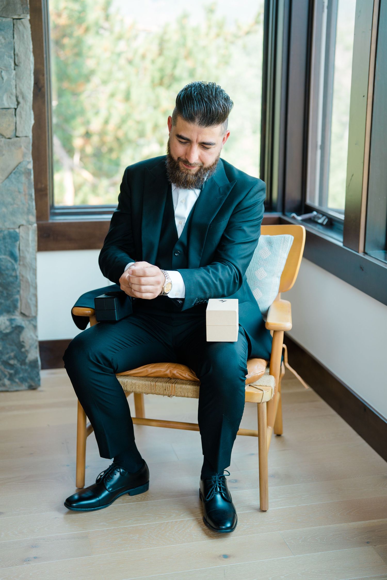 A man in a dark suit sits thoughtfully on a wooden chair by large windows in a sequence of four connected images during Breckenridge wedding