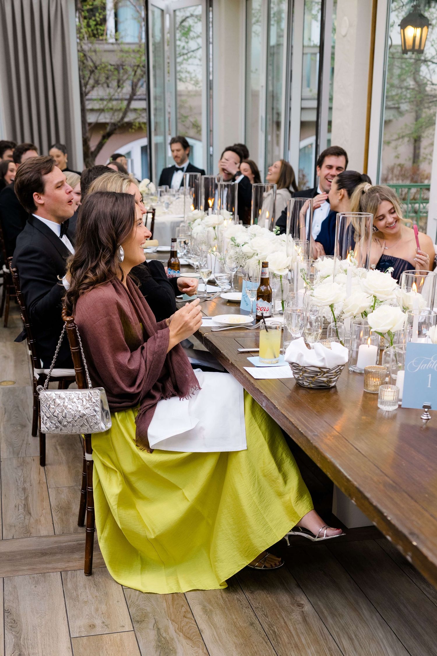 Guests laughing and clapping at an elegant wedding reception dinner with white floral centerpieces on a wooden table.
