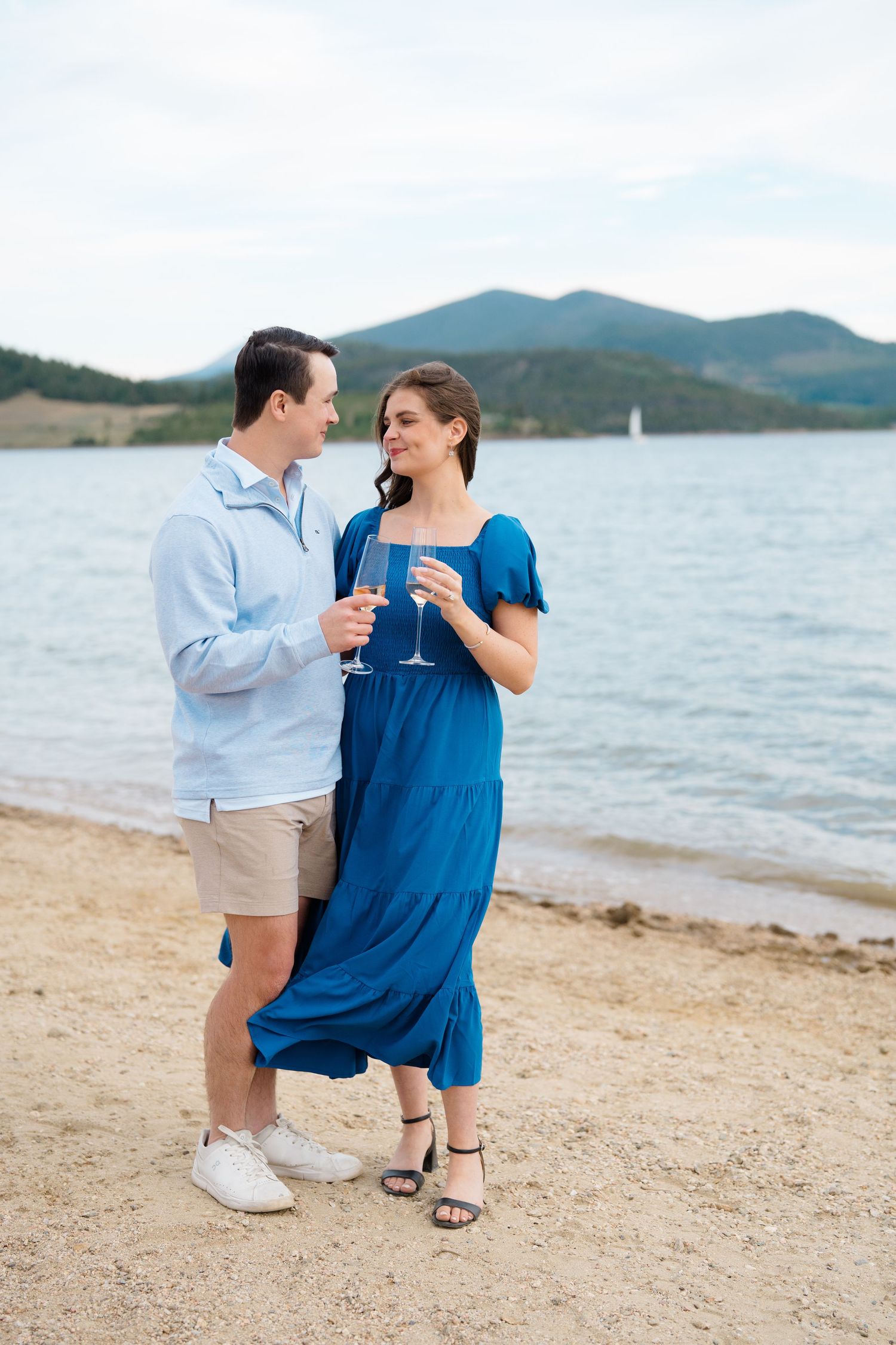 Couple sharing a romantic moment on a sandy beach with mountains and lake in the background.