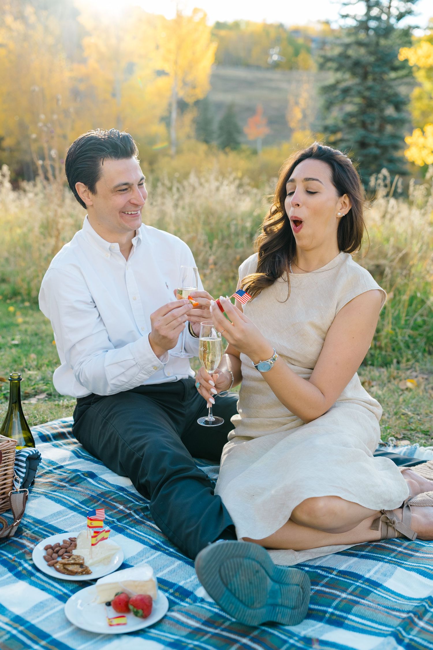 A couple enjoys a romantic autumn picnic on a blue blanket with wine and dessert in a golden outdoor setting.