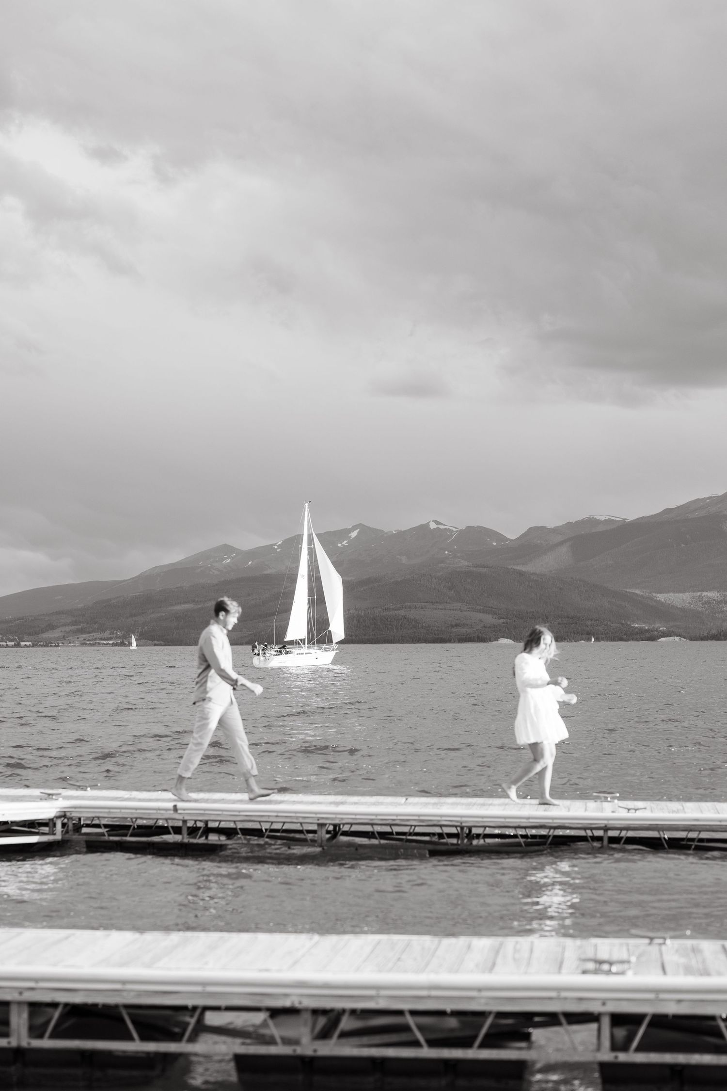 Black and white panoramic sequence showing silhouettes of people walking and jumping on a dock with sailboats in background.
