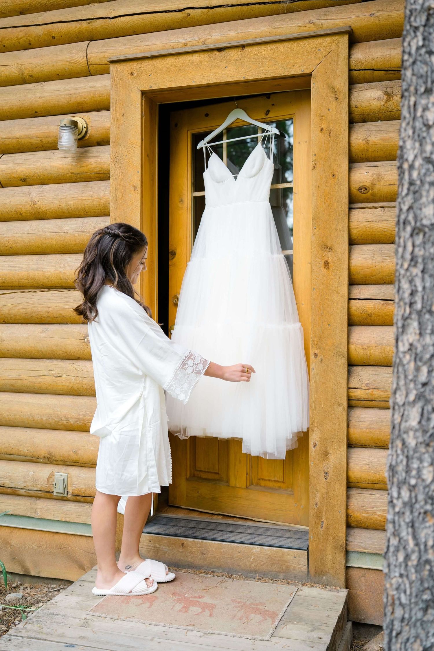 White Camp Hale wedding dress hangs on rustic log cabin exterior while someone in a white robe stands nearby.