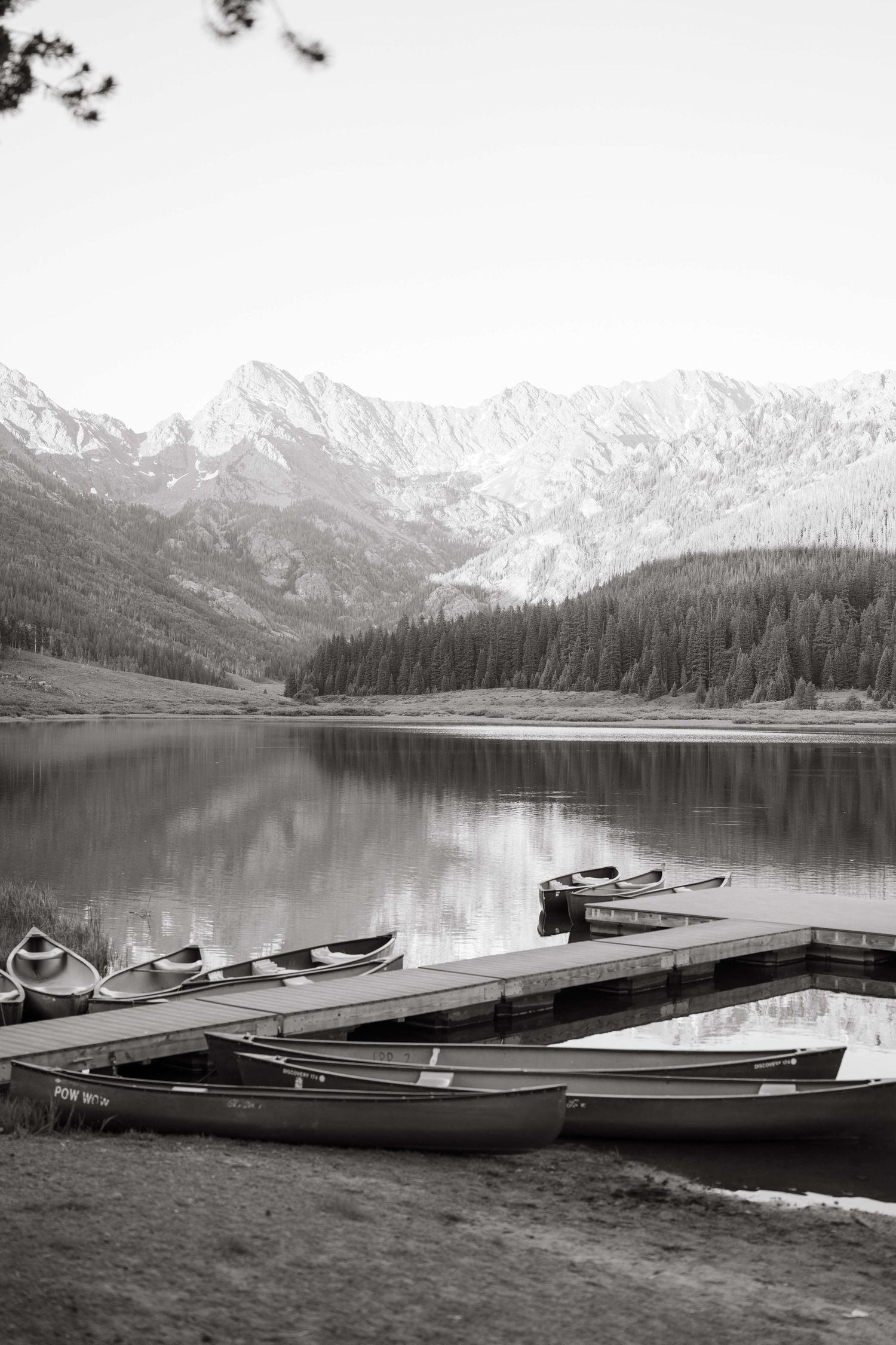 Black and white photo of canoes docked at a piney river ranch wedding with snow-capped peaks and pine forest reflected in calm water.