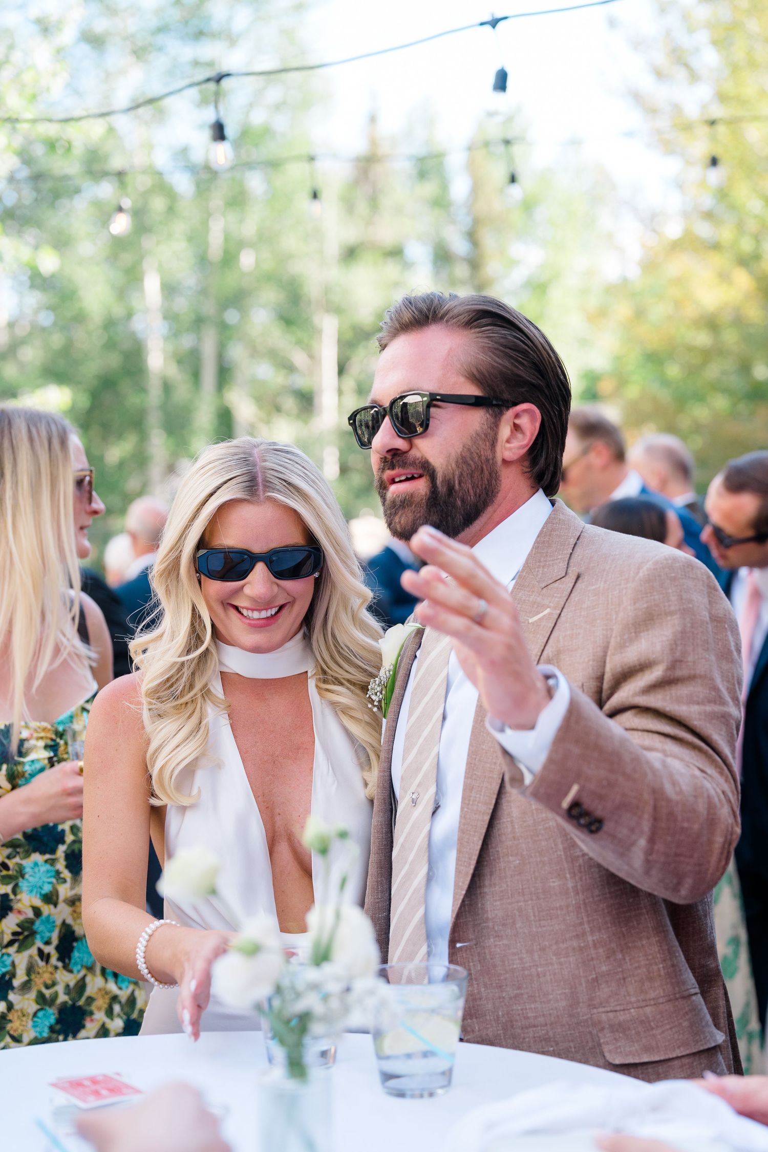 A blonde woman and bearded man in sunglasses chat at an outdoor wedding reception, dressed in elegant attire.