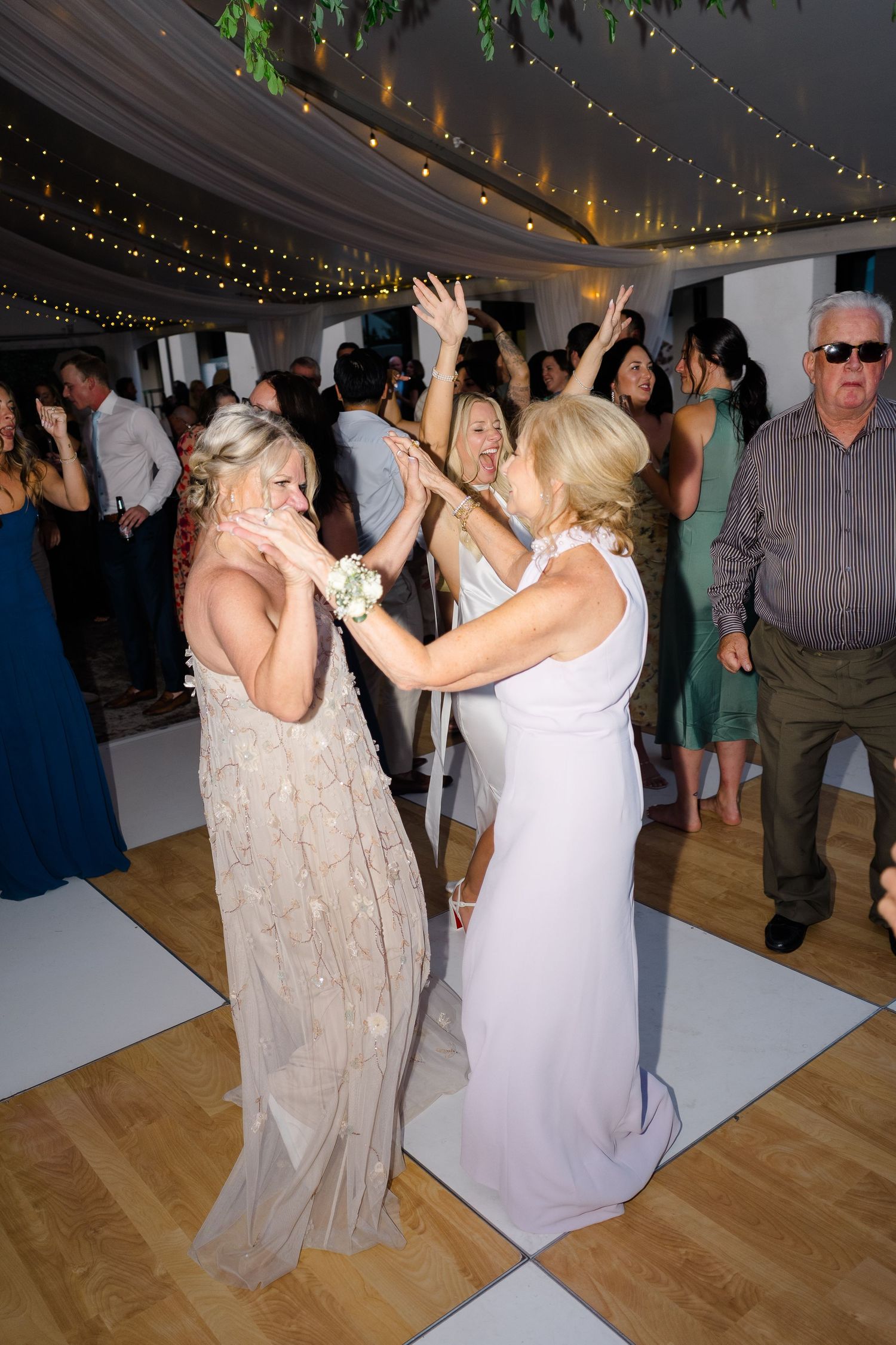 Women in elegant gowns dancing joyfully at a wedding reception under fairy lights on a white dance floor.