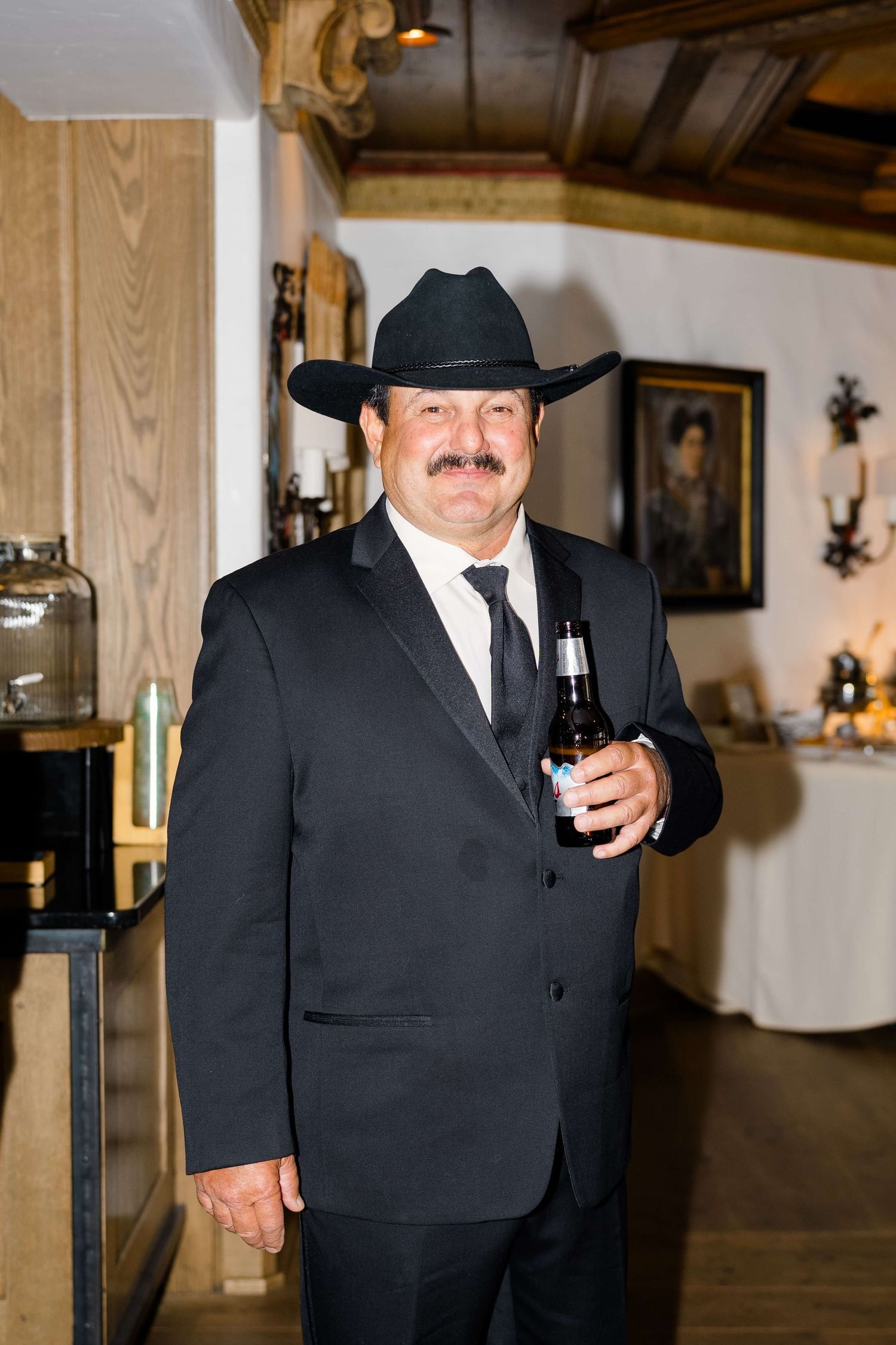 Man in black suit and cowboy hat holding a beer bottle at a rustic indoor wedding reception.