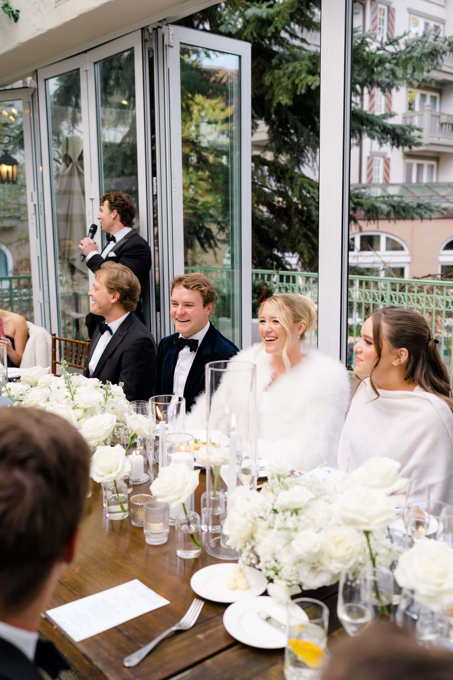 Bride laughing at head table with white floral arrangements during elegant wedding reception in glass venue.