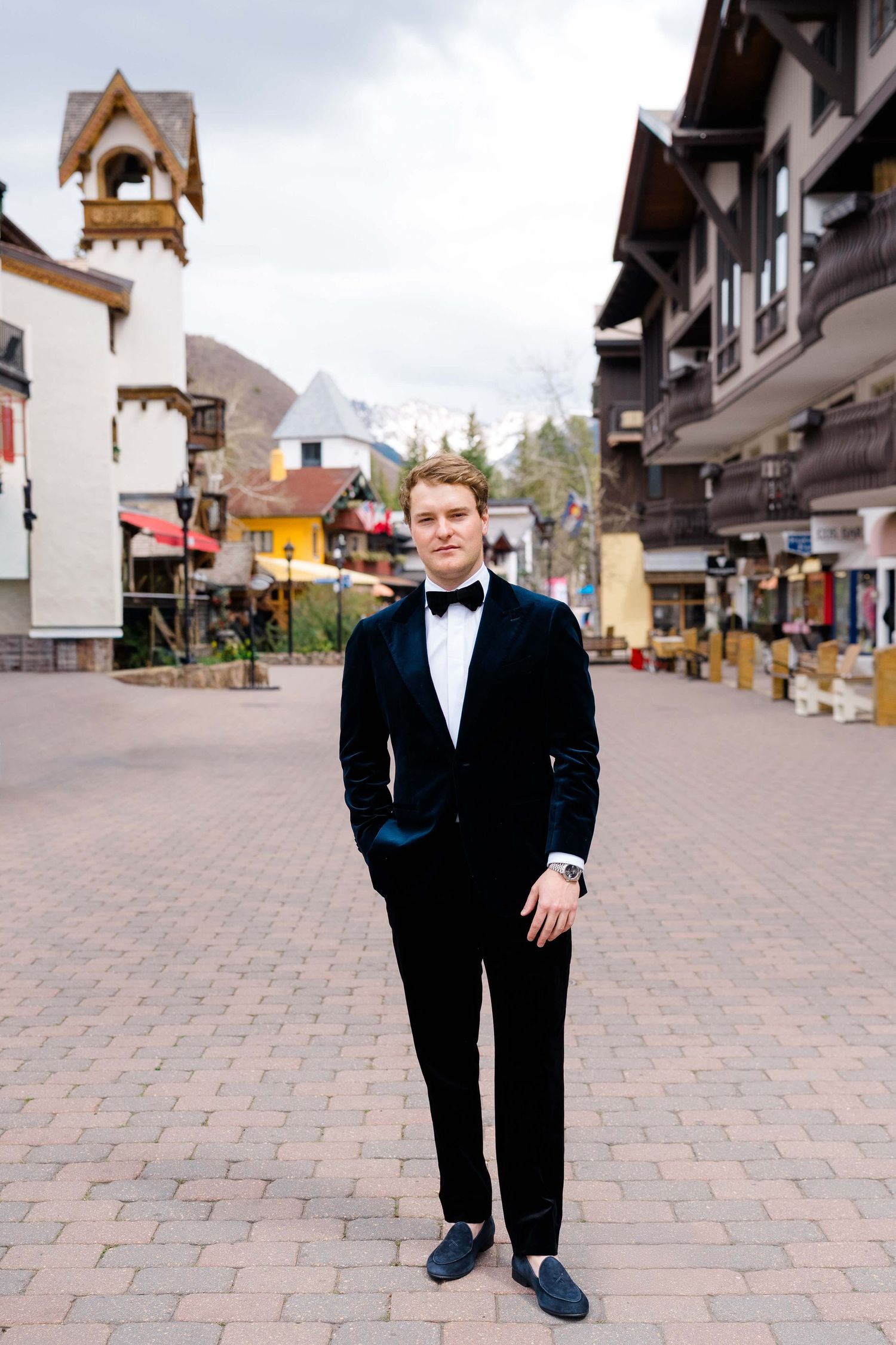 Young man in black tuxedo with bow tie standing in a European-style village square with alpine architecture.