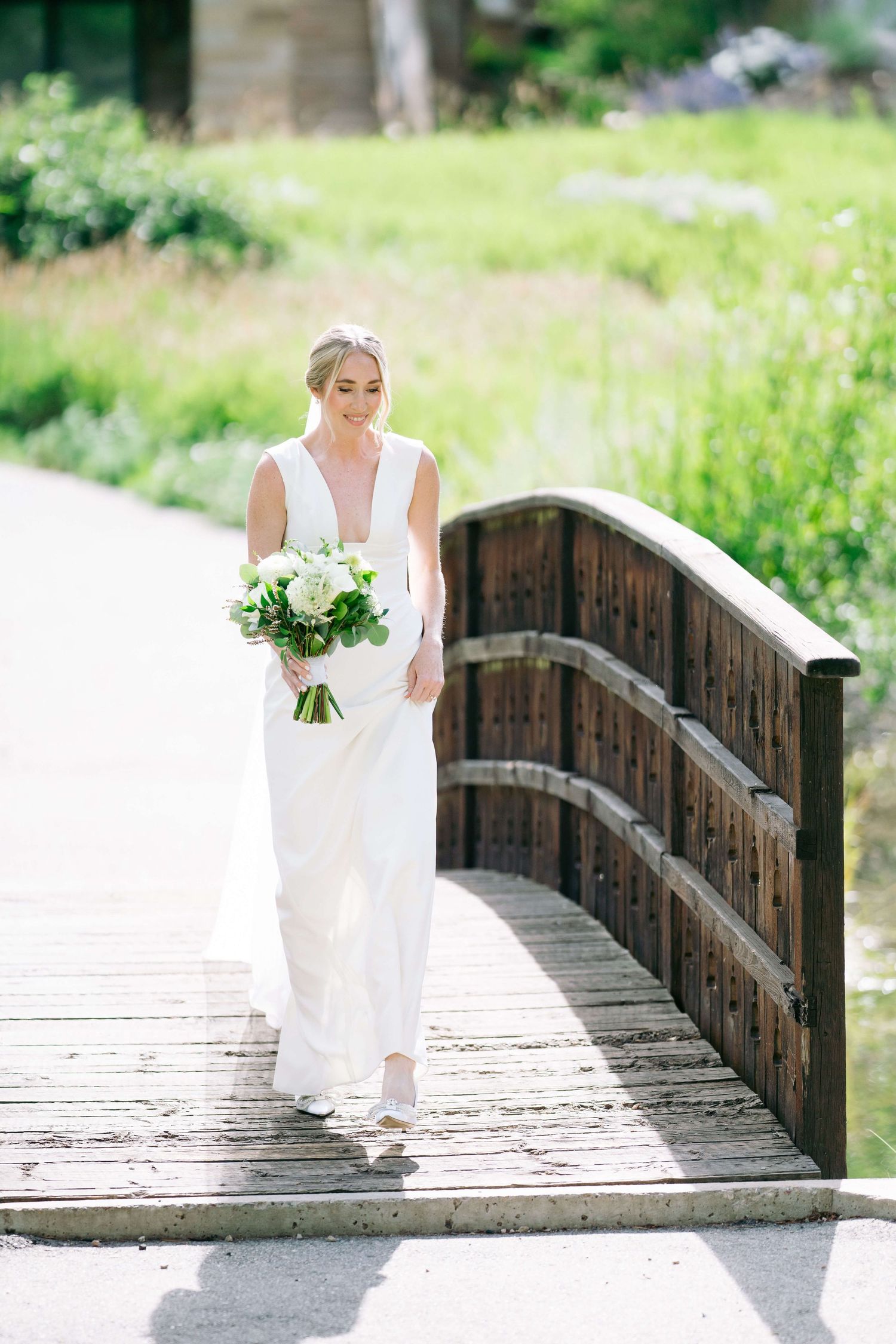 At Vail Golf Club Wedding Venue Bride in white gown holding bouquet walks on wooden bridge surrounded by lush greenery on sunny wedding day.