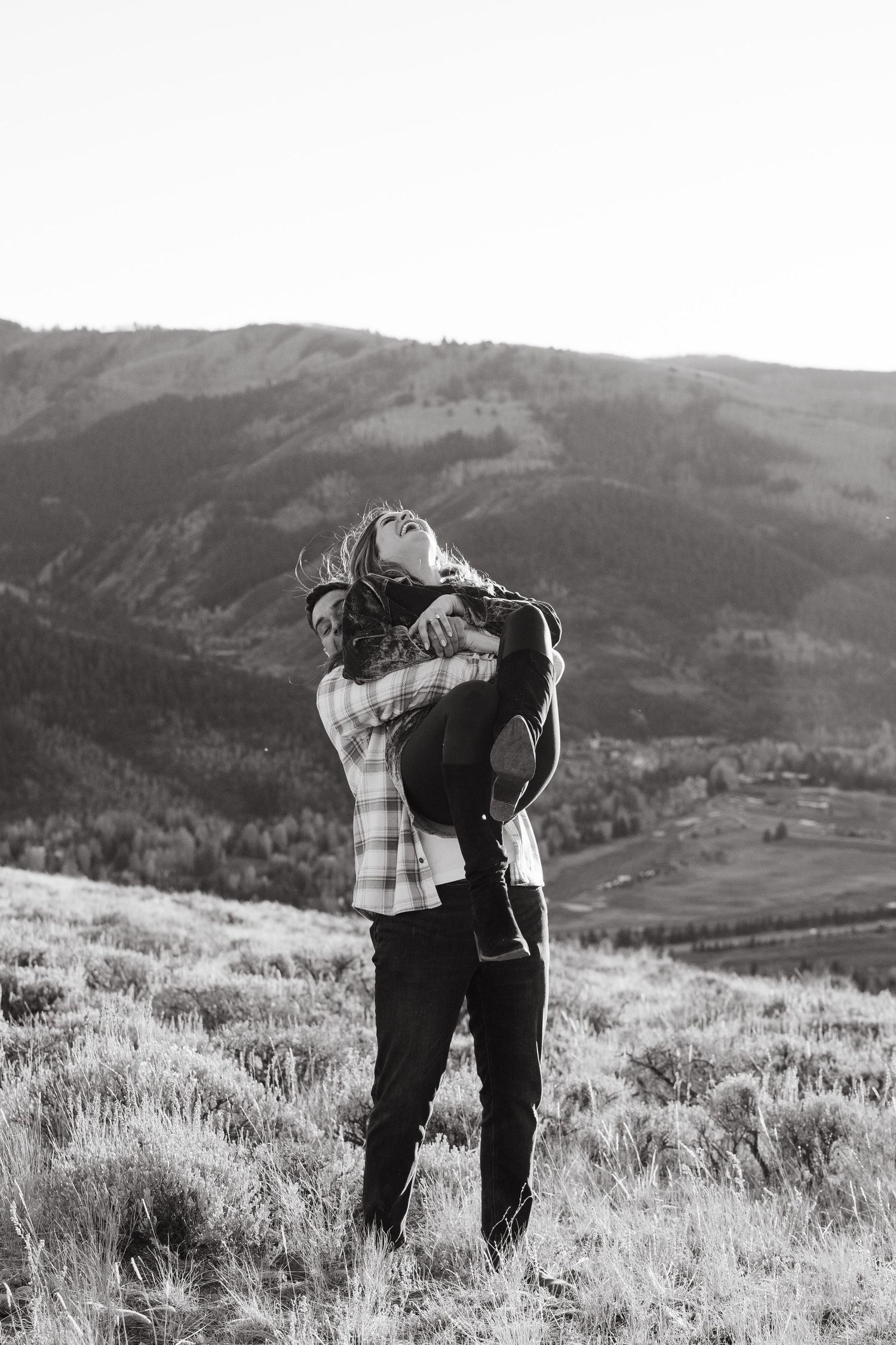 A romantic couple embraces while standing in a scenic mountain meadow with rolling hills in black and white.