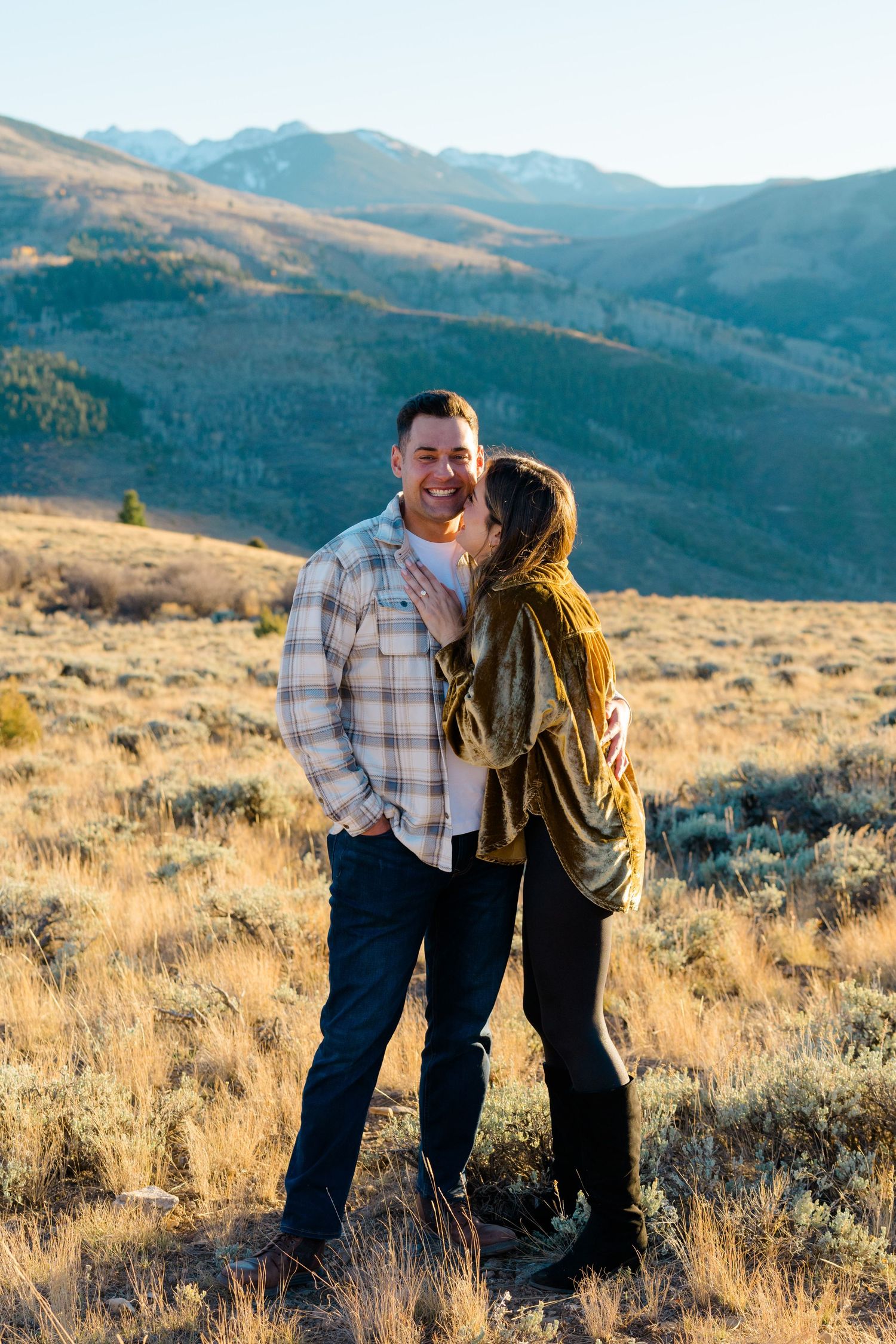 A couple shares a romantic moment in a golden mountain meadow at sunset with scenic mountain peaks in the background.