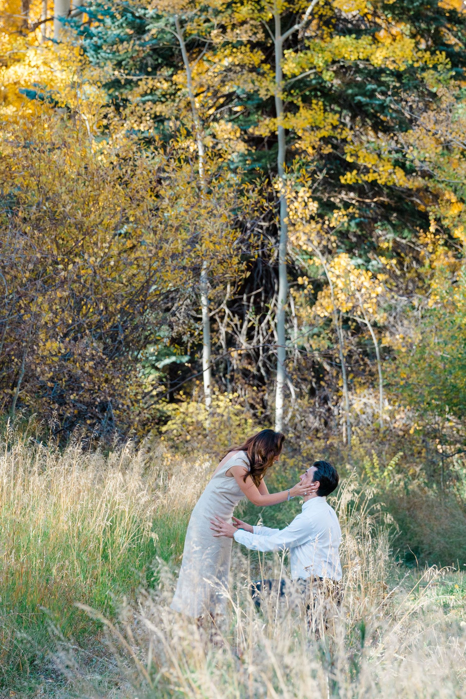 A couple shares a romantic moment in tall grass with autumn trees displaying golden foliage in the background.