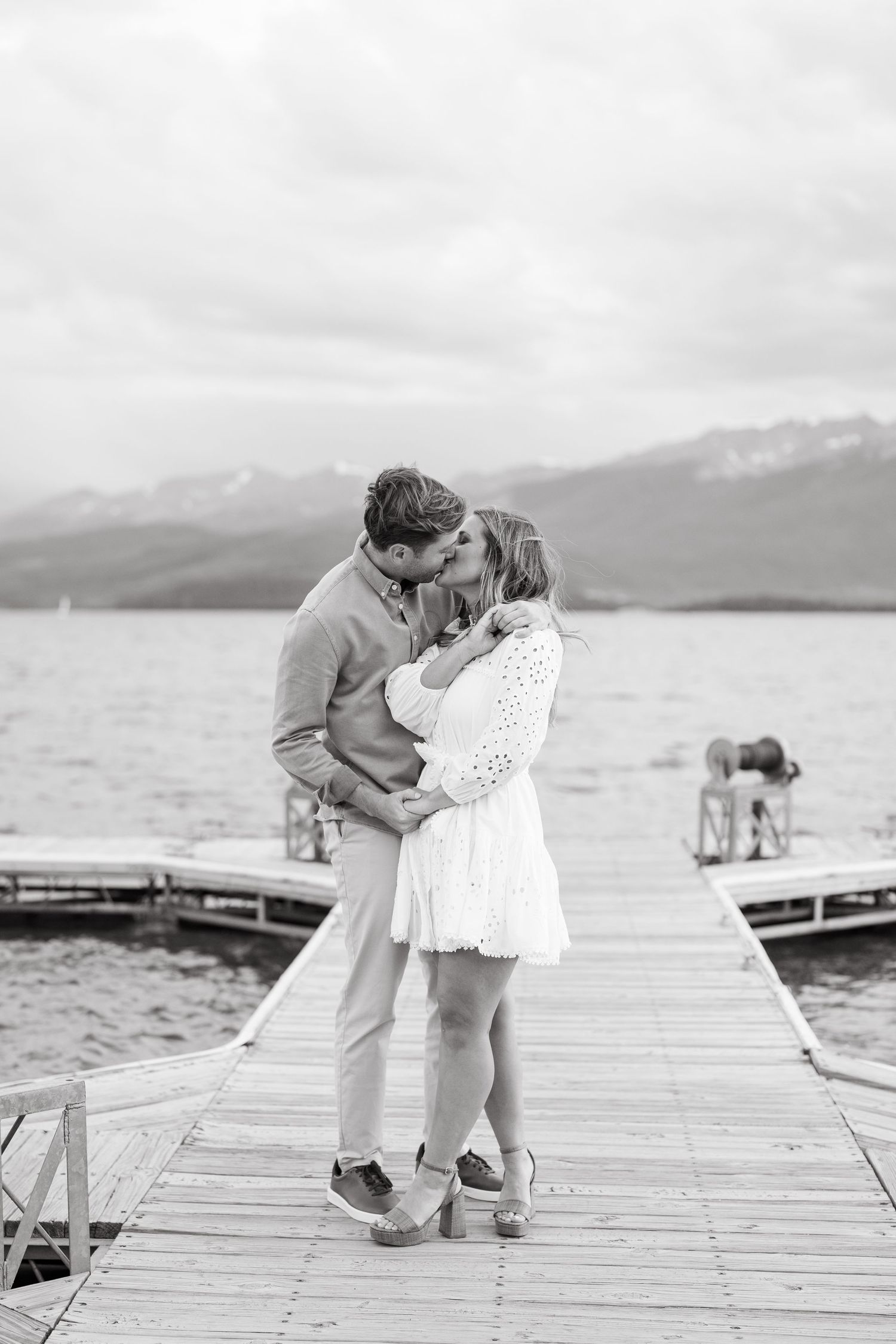 A romantic couple embraces on a wooden dock overlooking a serene mountain lake on an overcast day.