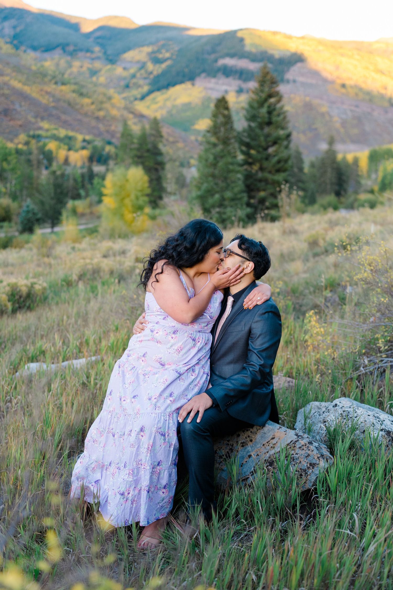 A romantic couple shares an intimate moment on a grassy hillside with mountains and evergreen trees in the background at sunset.