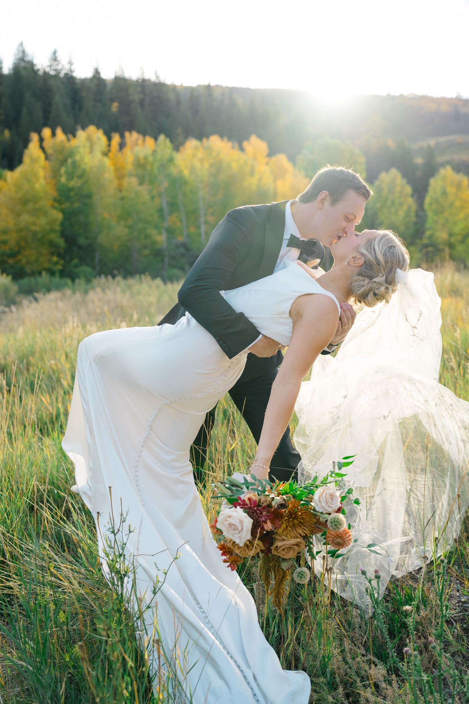 Wedding couple embraces in golden field during sunset with bride's veil flowing in the breeze.