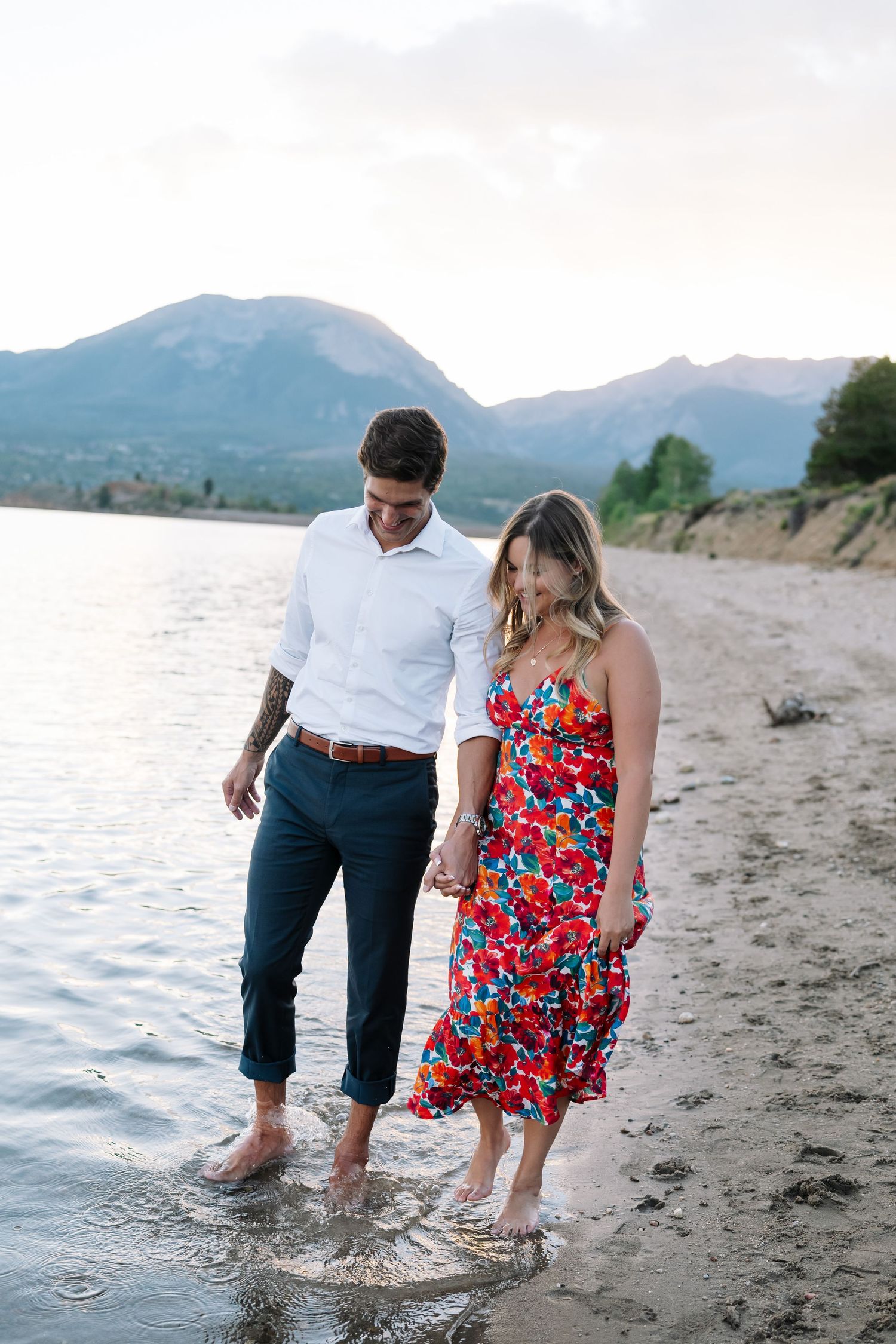 A couple wades together in shallow water along a scenic mountain beach at sunset.
