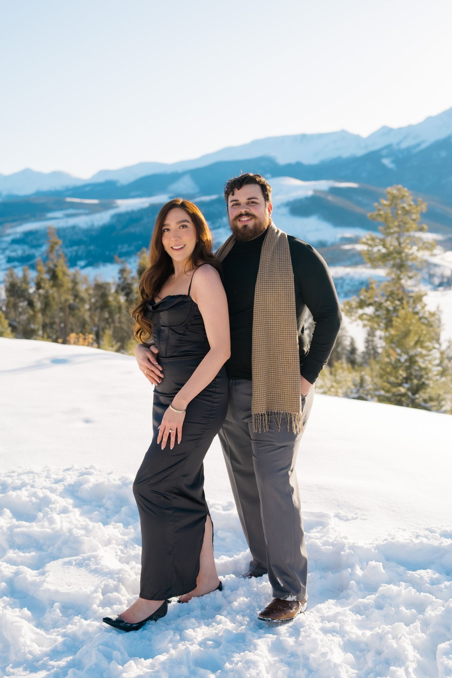 A couple poses together in formal attire on a snowy mountain overlook with evergreen trees and peaks in the background.