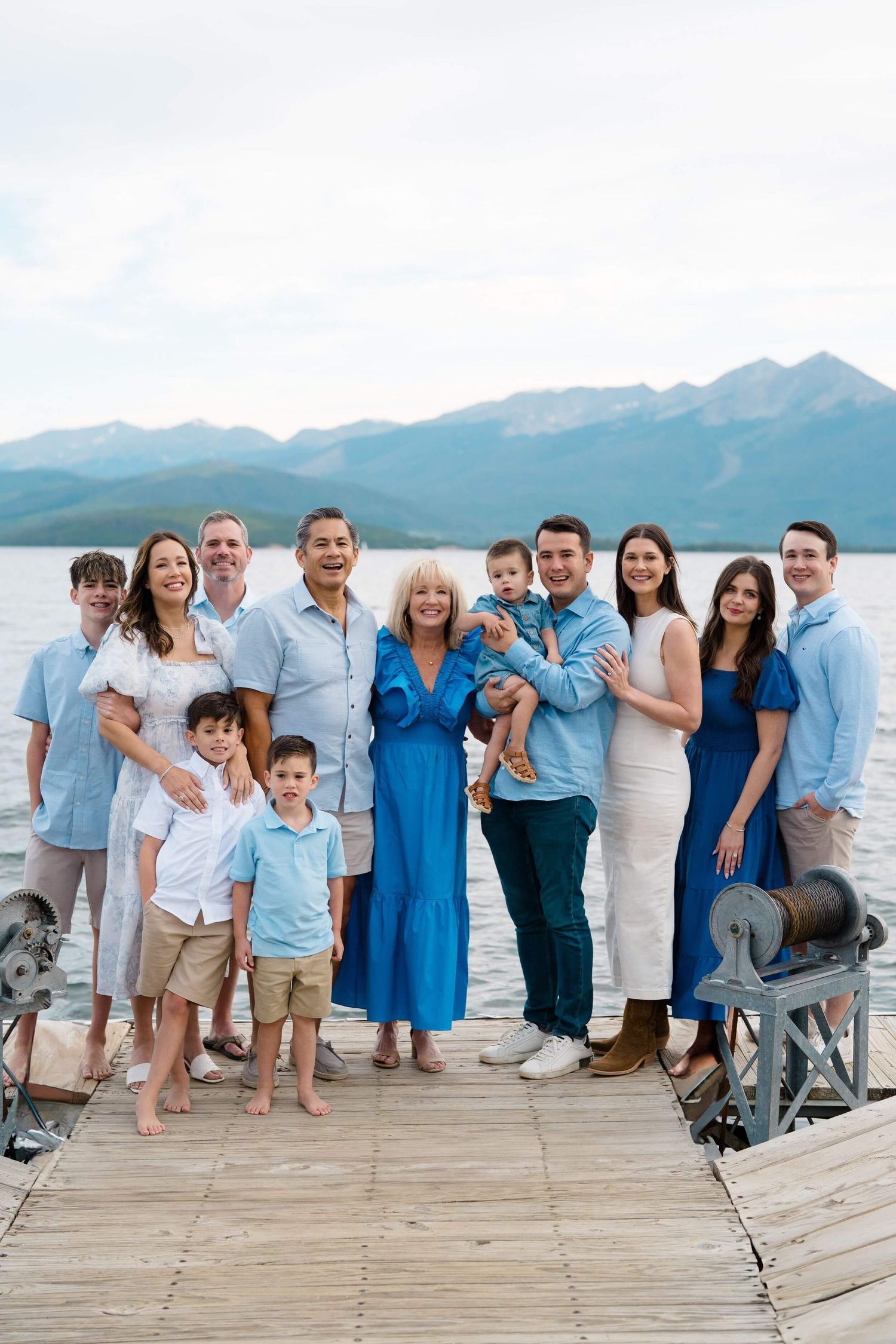 Extended family group poses together on a wooden dock with scenic mountain lake backdrop in coordinating blue and white outfits.