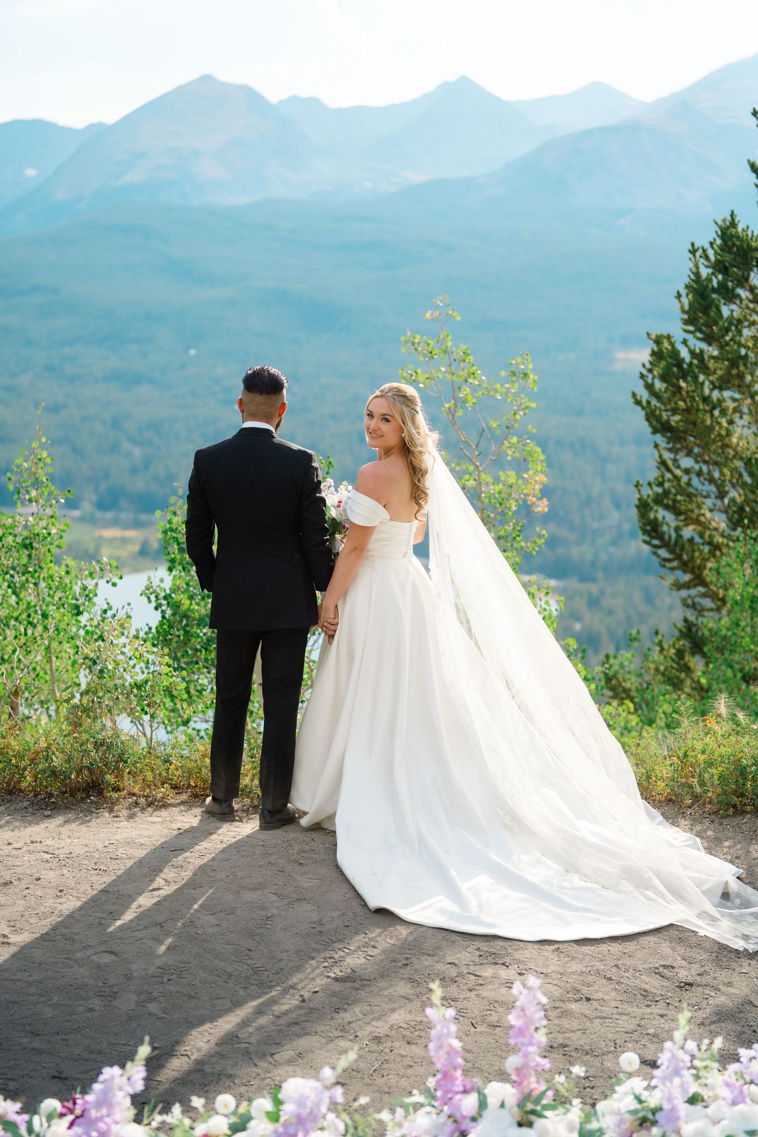 A couple in wedding attire stands at a scenic vail mountain overlook with a dramatic mountain landscape in the background.
