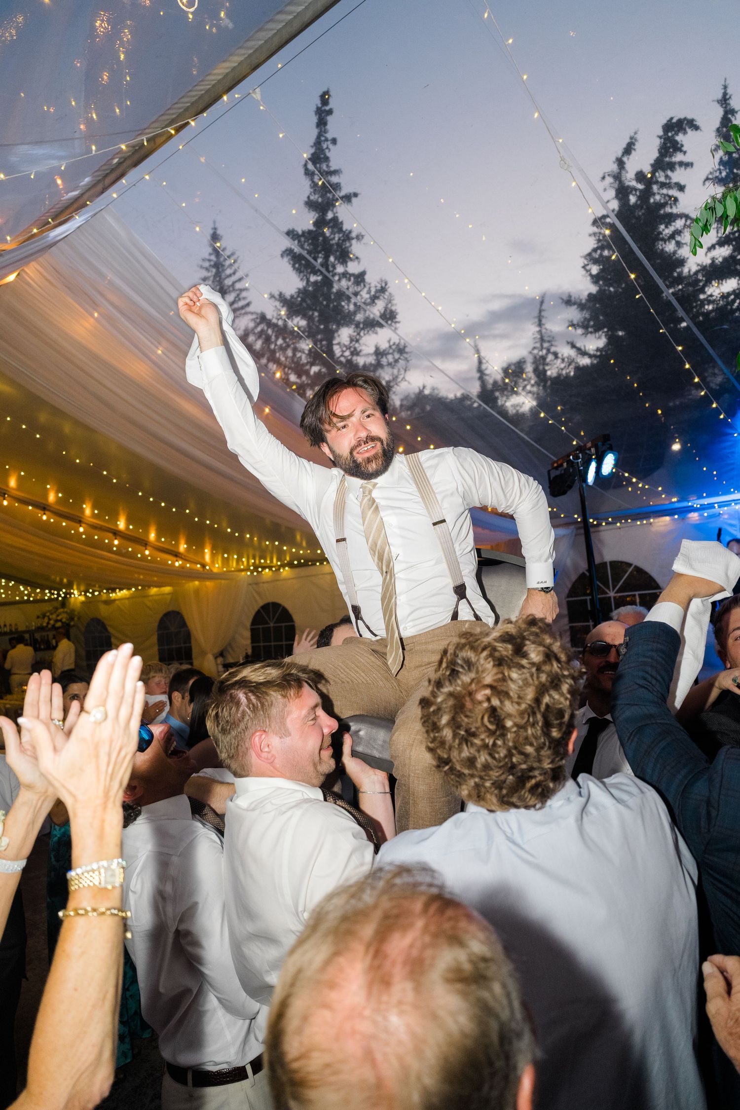 Groom lifted by guests at wedding reception under fairy light tent, raising fist in celebration.