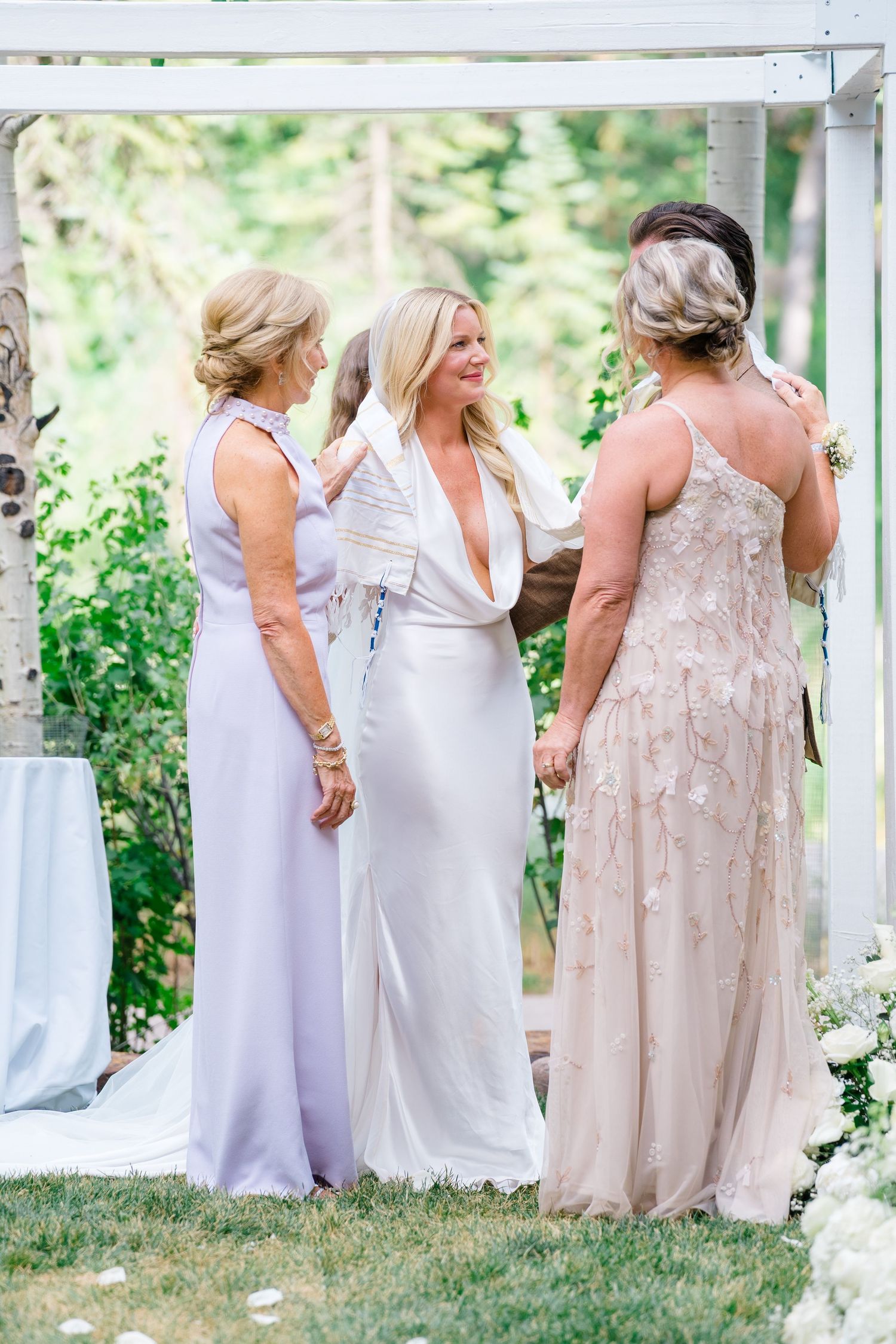 Bride in white gown stands with two women in lavender and blush dresses at an outdoor wedding ceremony.