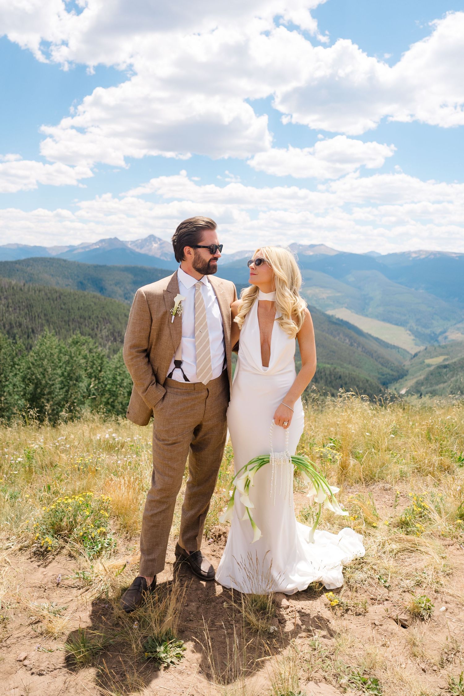 A bride and groom pose on a mountain hilltop in Colorado, she in a white halter gown, he in a tan suit with sunglasses.