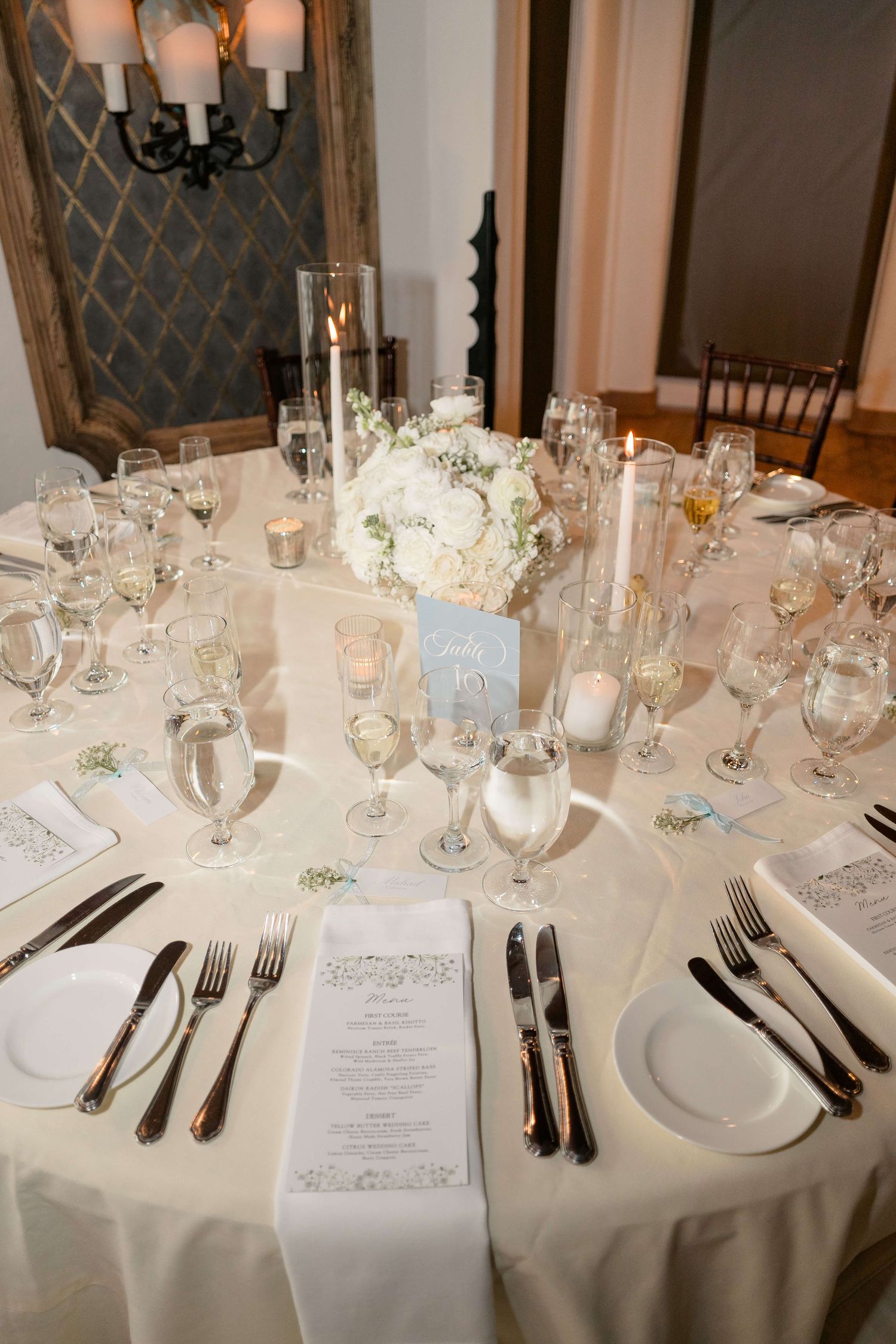Elegant wedding reception table set with white linens, crystal glassware, white floral centerpiece, and candles.