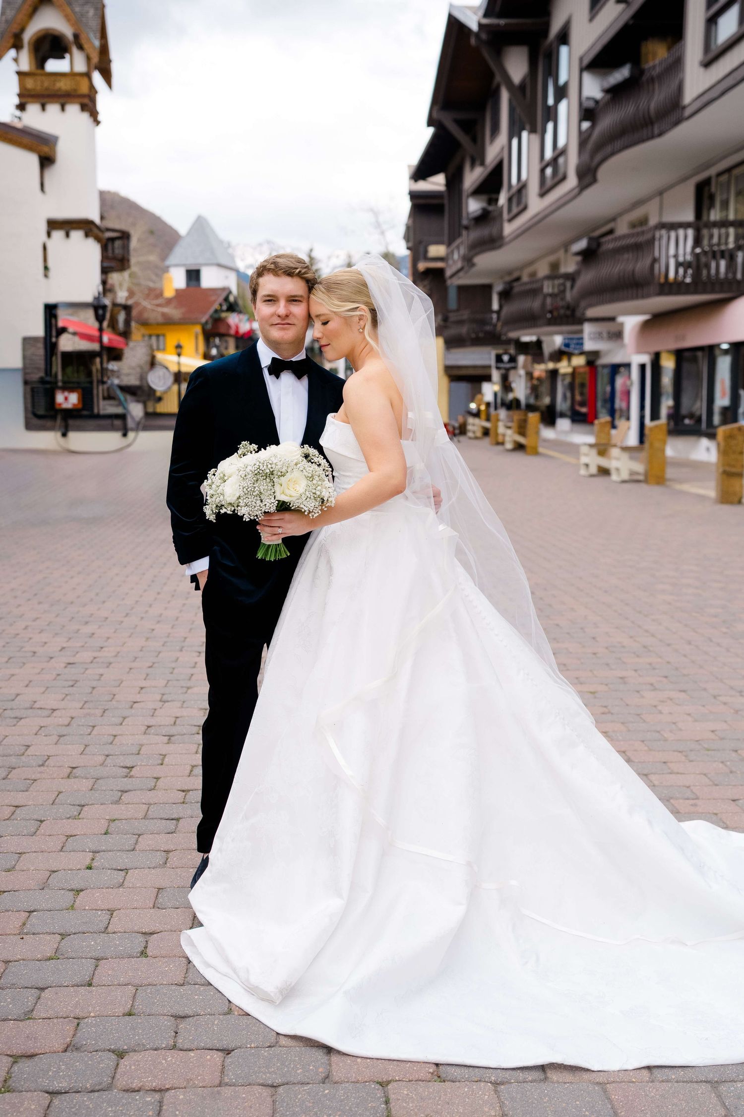 Bride in white ballgown with veil holds bouquet beside groom in black tuxedo on cobblestone street.
