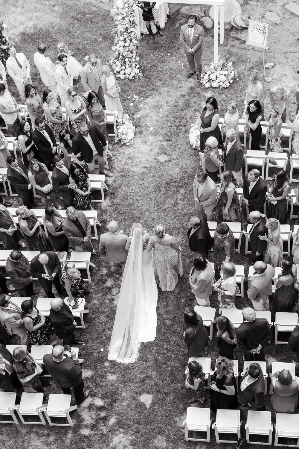 Aerial black and white photo of a bride walking down the aisle at an outdoor wedding ceremony.