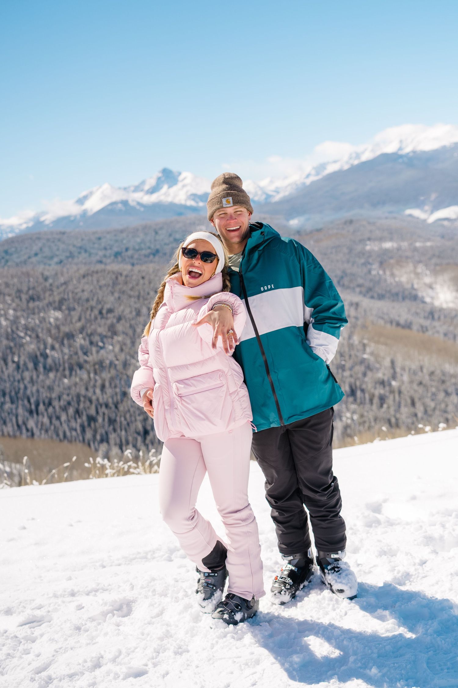 Two people share a playful moment in the snow with snow-capped mountains and evergreen forest in the background.